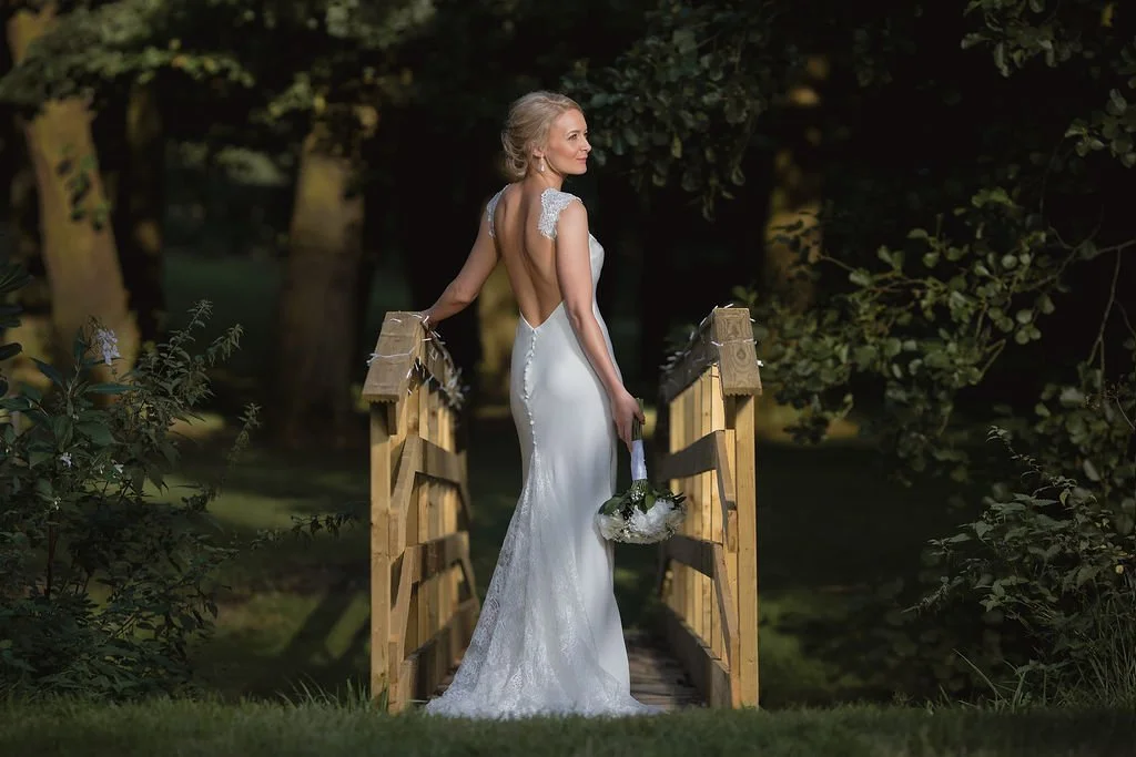 Bride standing on a small wooden bridge in an outdoor setting during the evening, wearing a backless white wedding gown and holding a bouquet.