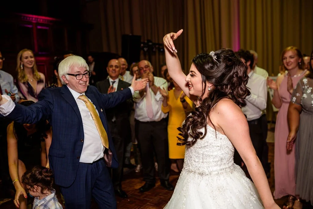 A bride and an older man dancing at a wedding reception with guests clapping and smiling in the background.