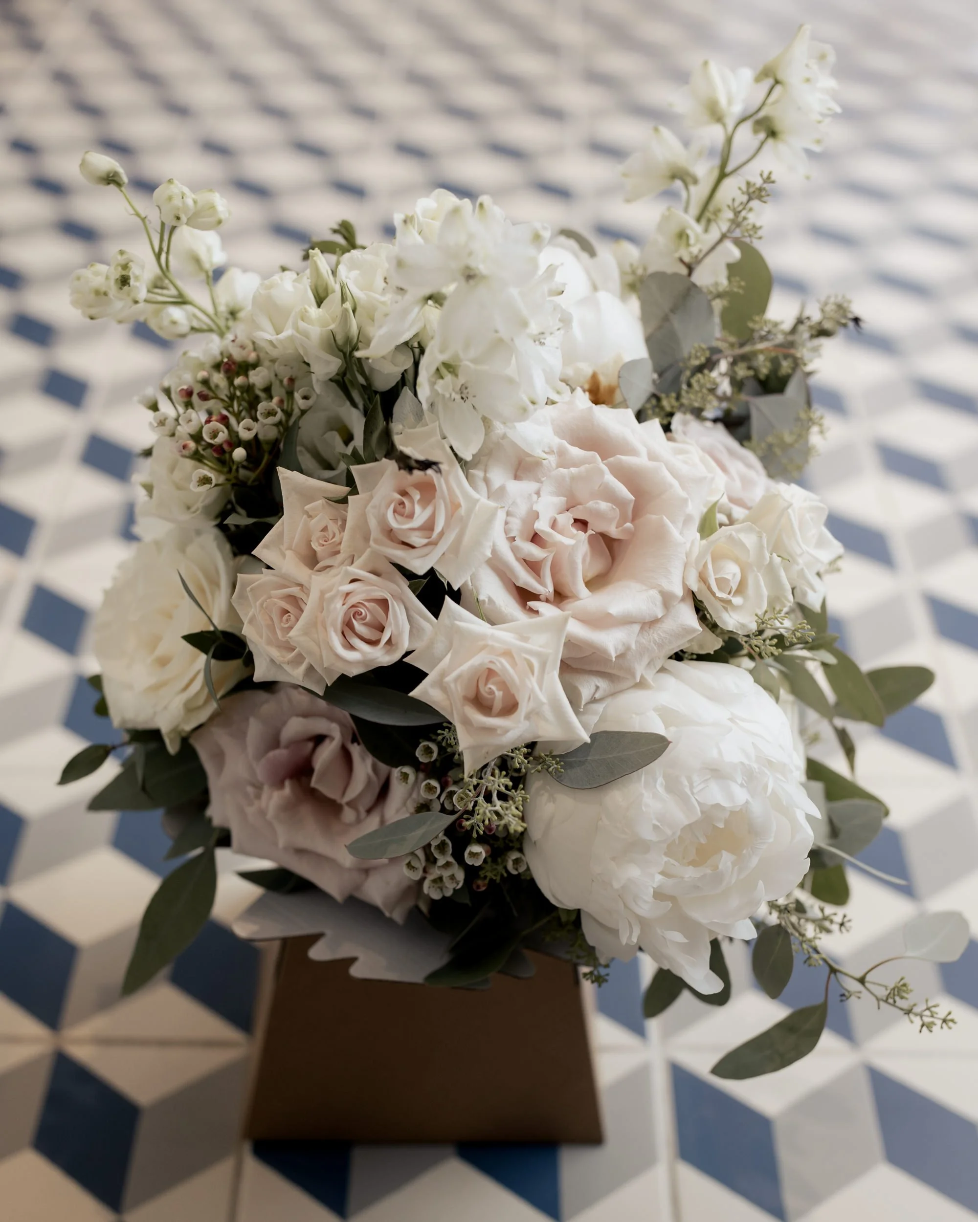 A bouquet of white and pale pink flowers, including roses and other blossoms, with green and gray foliage, placed on a wooden surface. The background features a black and white checkered pattern.