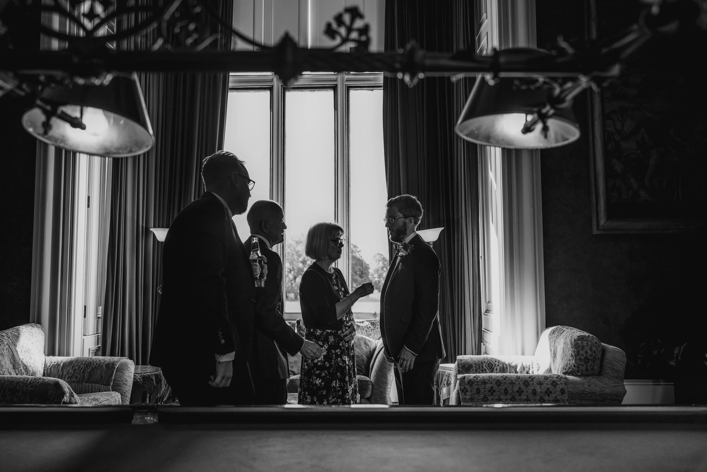 Black and white photo of five people standing indoors near a large window with curtains, engaged in conversation. The scene is backlit, casting silhouettes of the individuals, with sofas and wall lamps visible in the room.