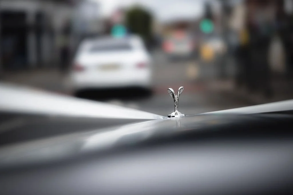 Close-up of a silver hood ornament on a luxury car, with a blurred city street in the background.