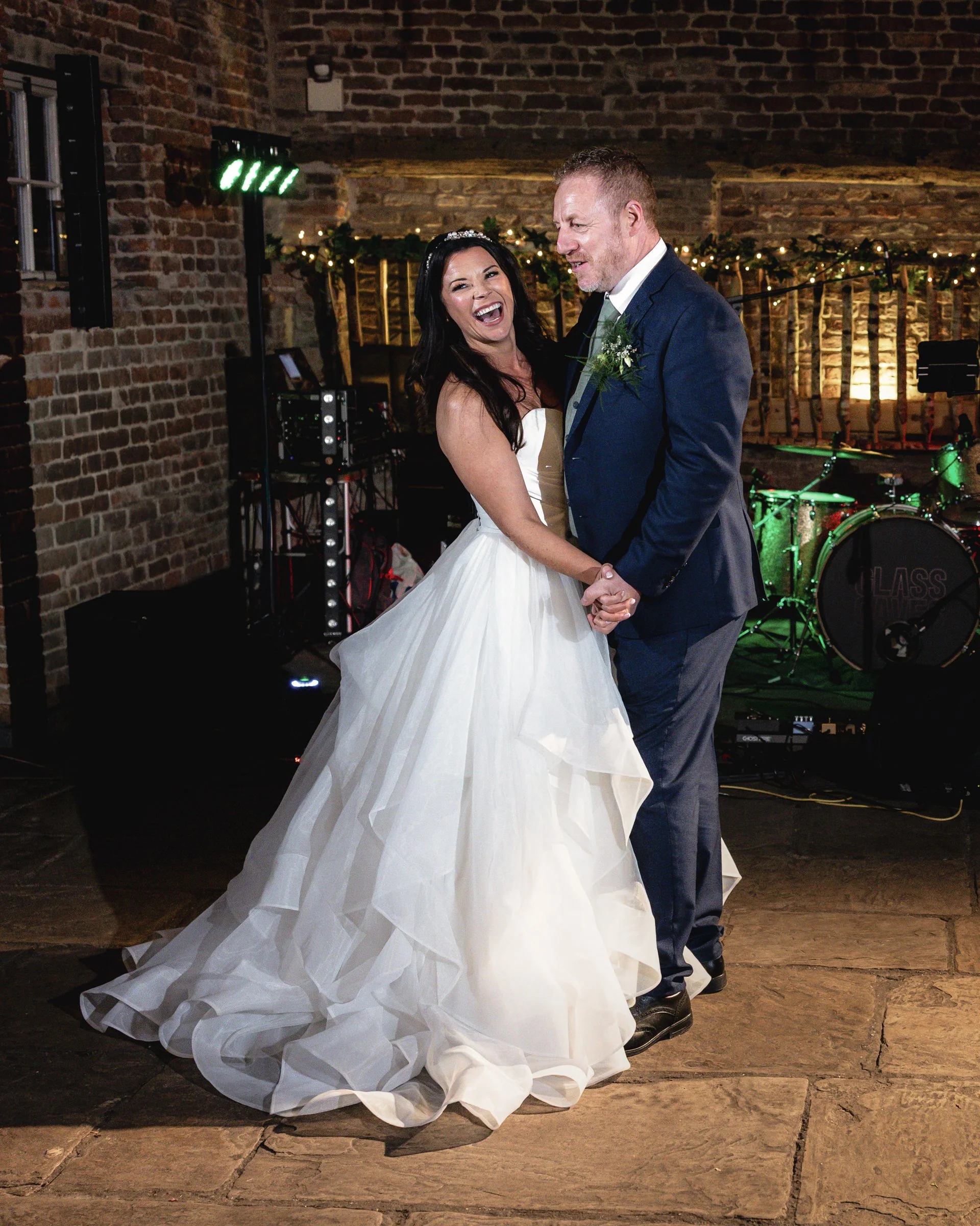 A bride and groom dancing at their wedding reception, sharing a joyful moment, with a brick wall, string lights, and a live band in the background.