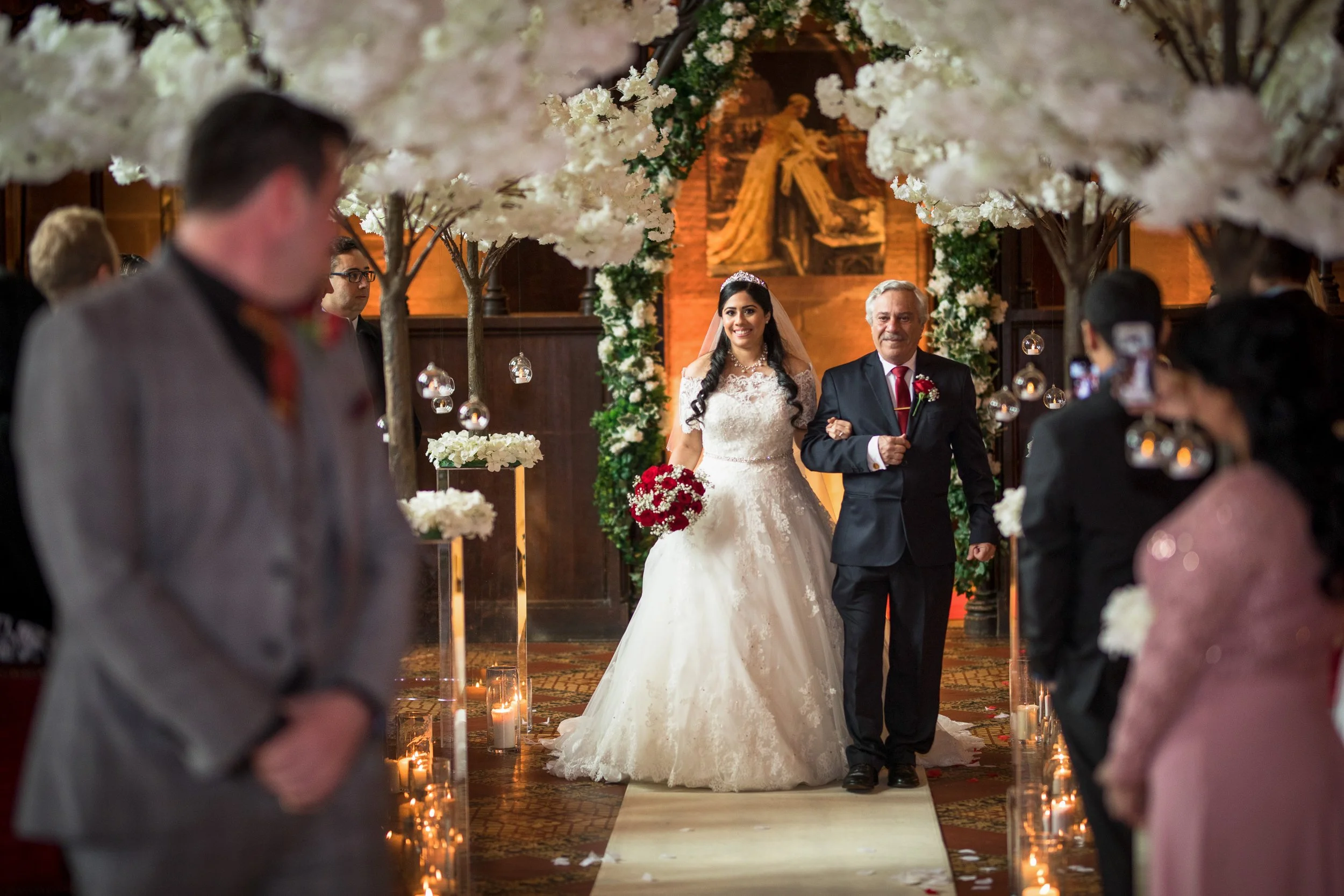 A bride in a white wedding gown walking down the aisle with her father during a wedding ceremony, surrounded by floral decorations and guests.