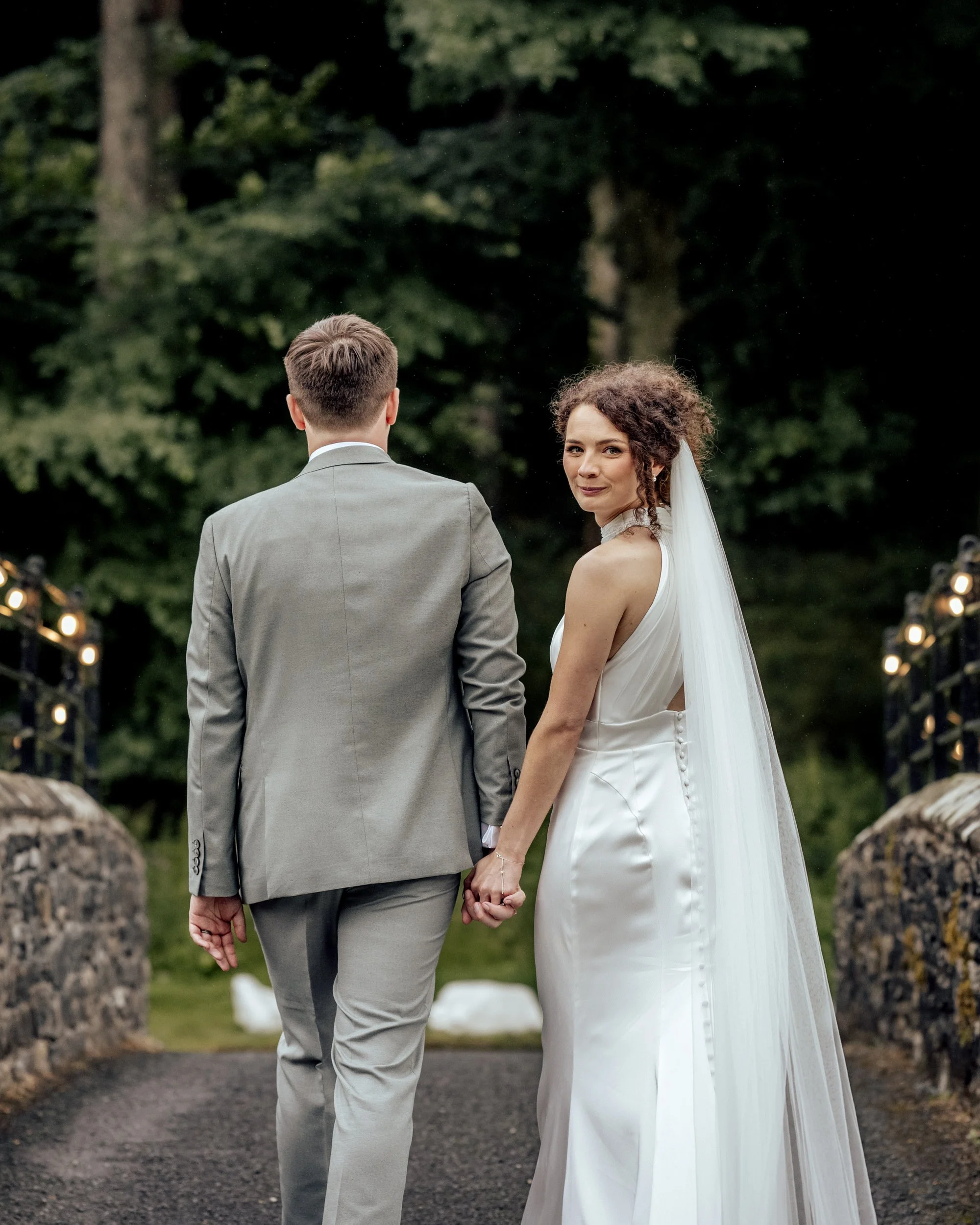 A bride and groom holding hands while walking outdoors, with the bride looking back and smiling, during a wedding ceremony in a natural setting with trees and string lights.