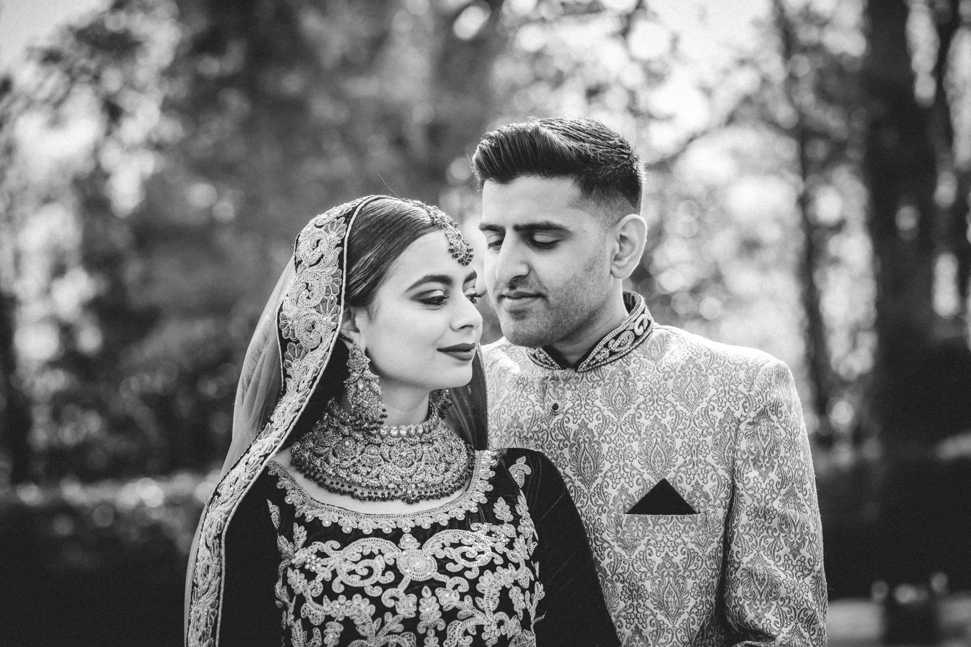 A bride and groom in traditional Indian wedding attire, looking at each other outdoors.