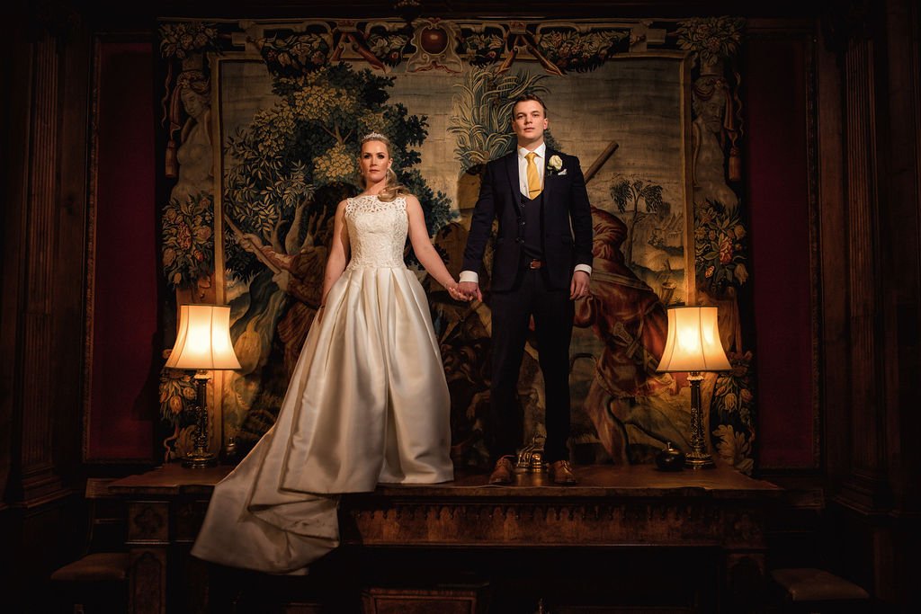 A bride and groom holding hands on a small stage, with vintage lamps on each side, in front of a tapestry depicting a landscape scene, with dark wood paneling on the walls.
