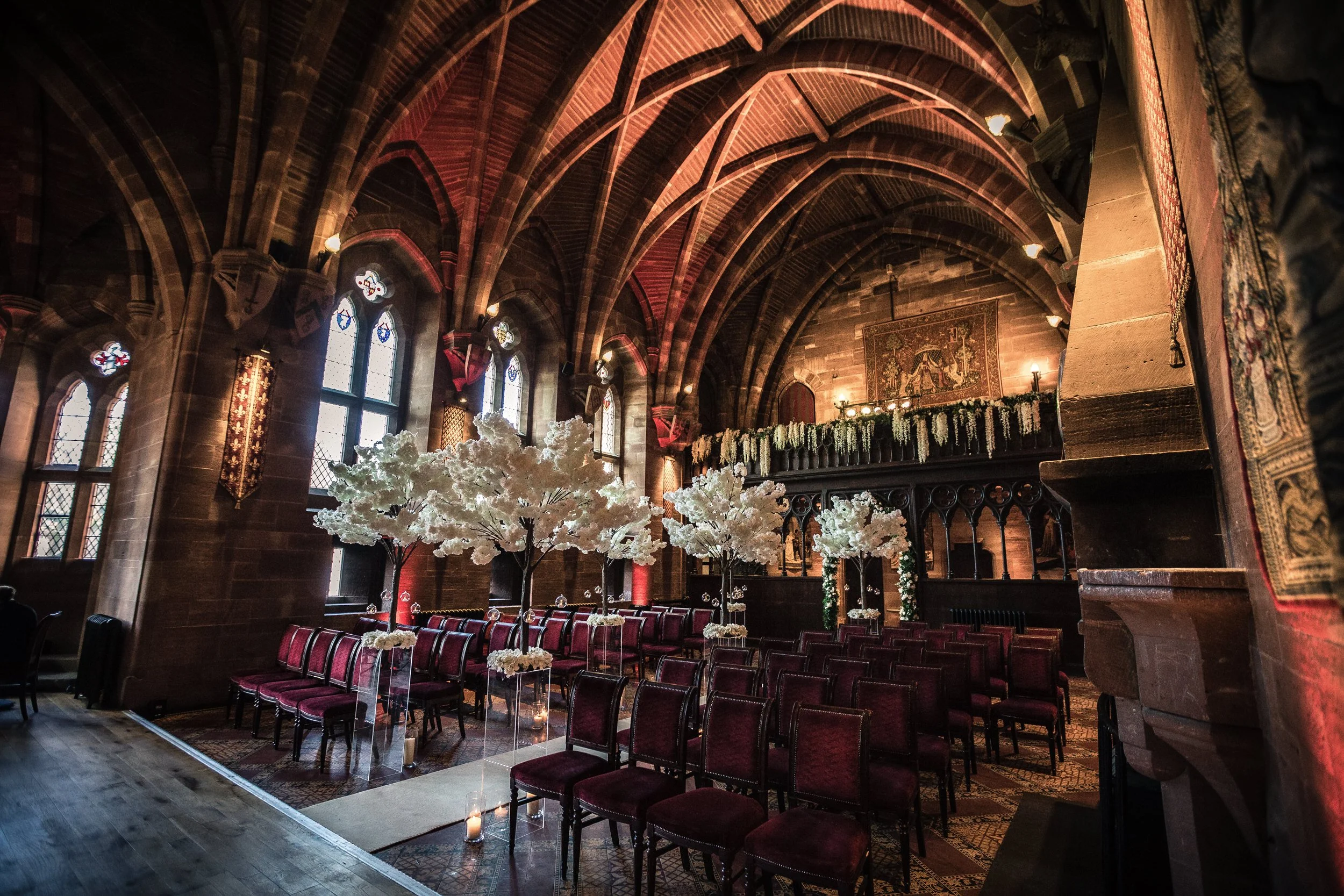 Inside of a gothic-style church decorated for a wedding, with white floral trees, red velvet chairs, and hanging floral arrangements