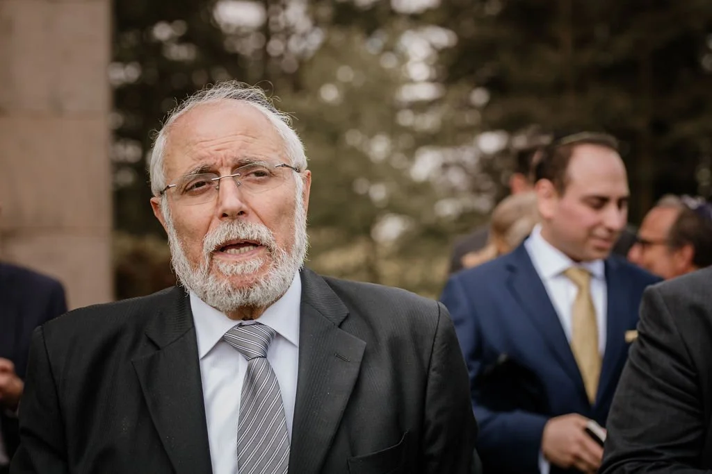 An elderly man with glasses and a beard in a black suit and striped tie, talking outdoors at a formal gathering.