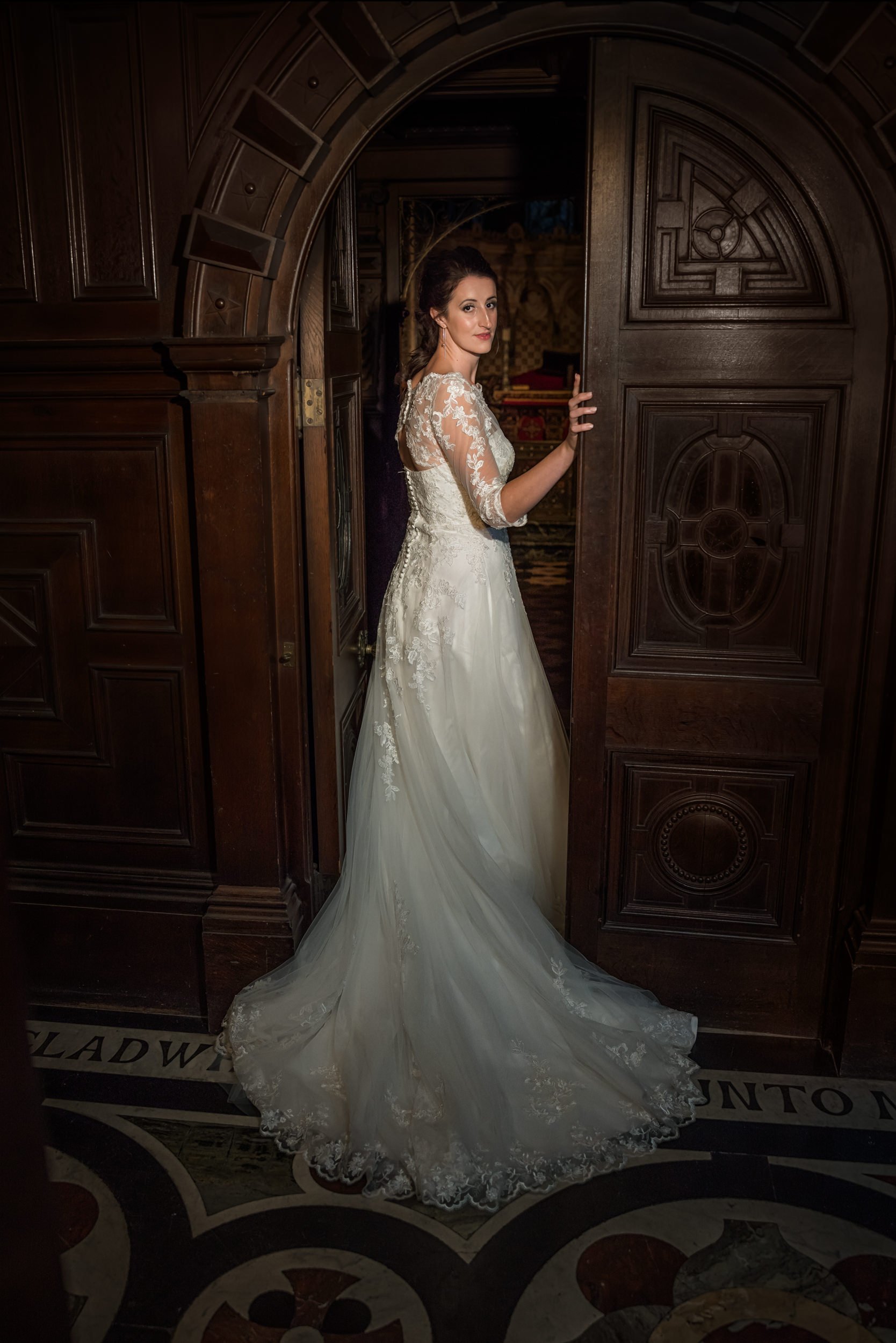 A woman in a white wedding dress with lace details, standing partially behind a large dark wooden door in a historic, ornate room, looking at the camera.