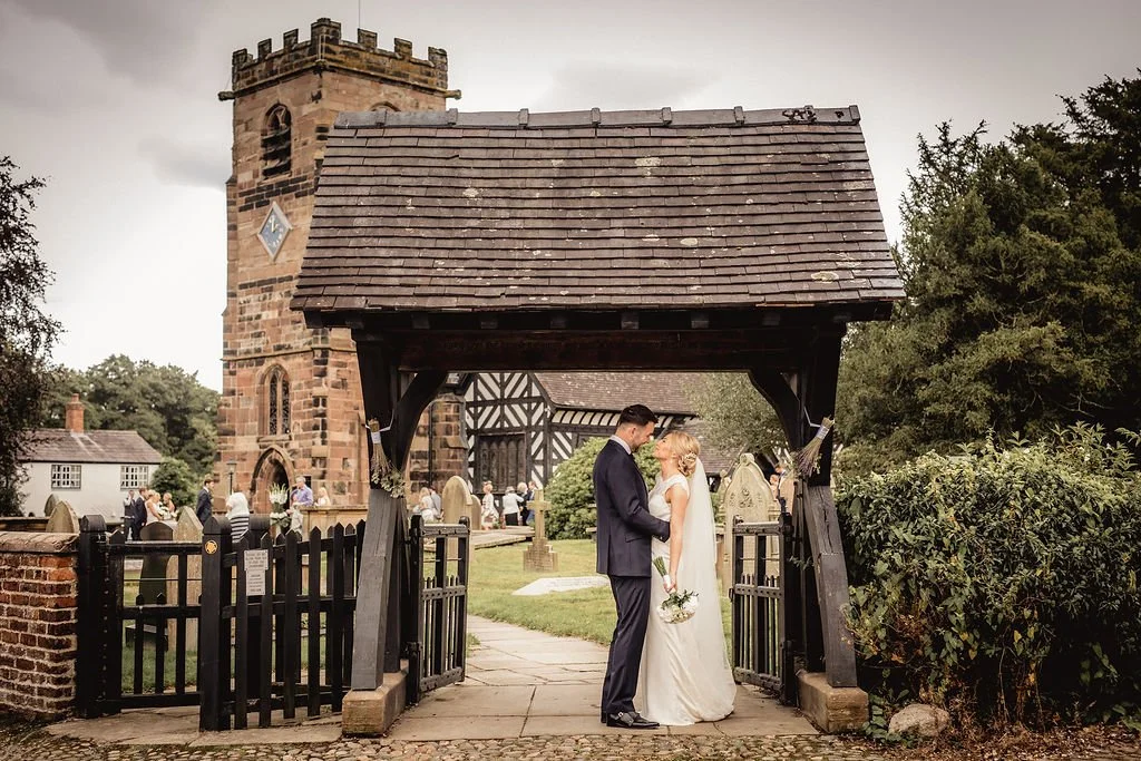 A bride and groom holding hands and facing each other under a small wooden gate, with a church and wedding guests in the background during a wedding ceremony.