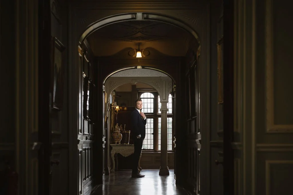 A man in a suit standing in a richly decorated hallway with wood paneling, ornate furniture, and large windows.