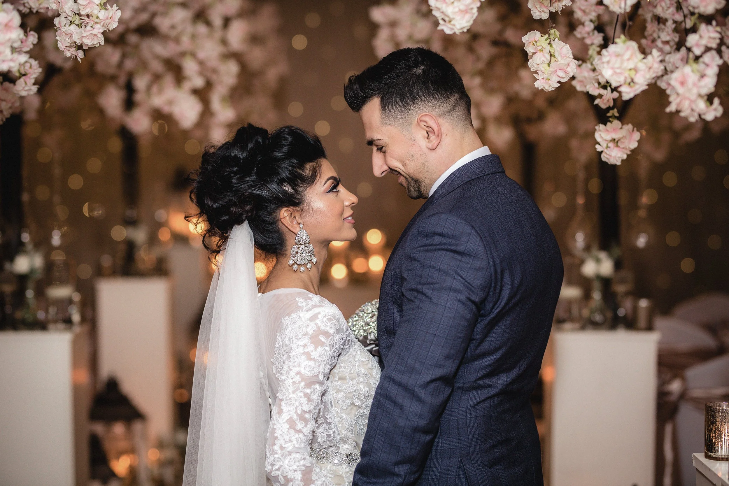 A bride and groom face each other smiling at their wedding reception, with pink flowers hanging above and blurred lights in the background.