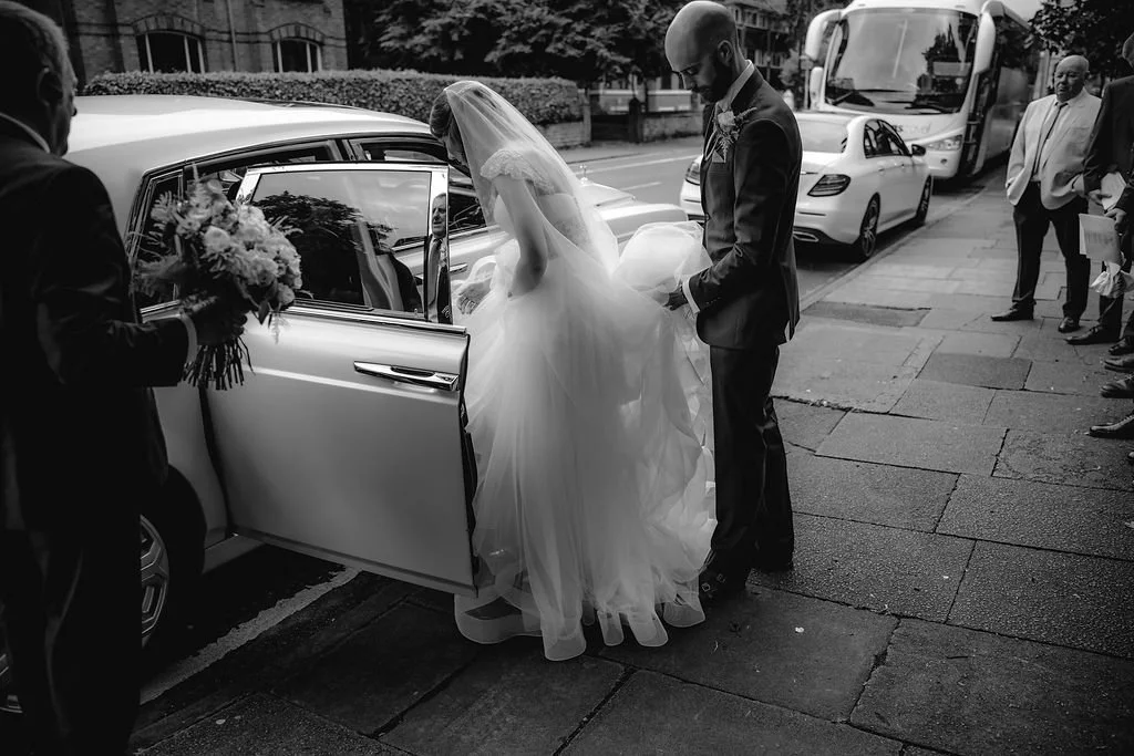 A bride in a wedding dress getting into a car with a groom helping her, while a person on the left holds a bouquet of flowers. Several onlookers and vehicles are in the background.