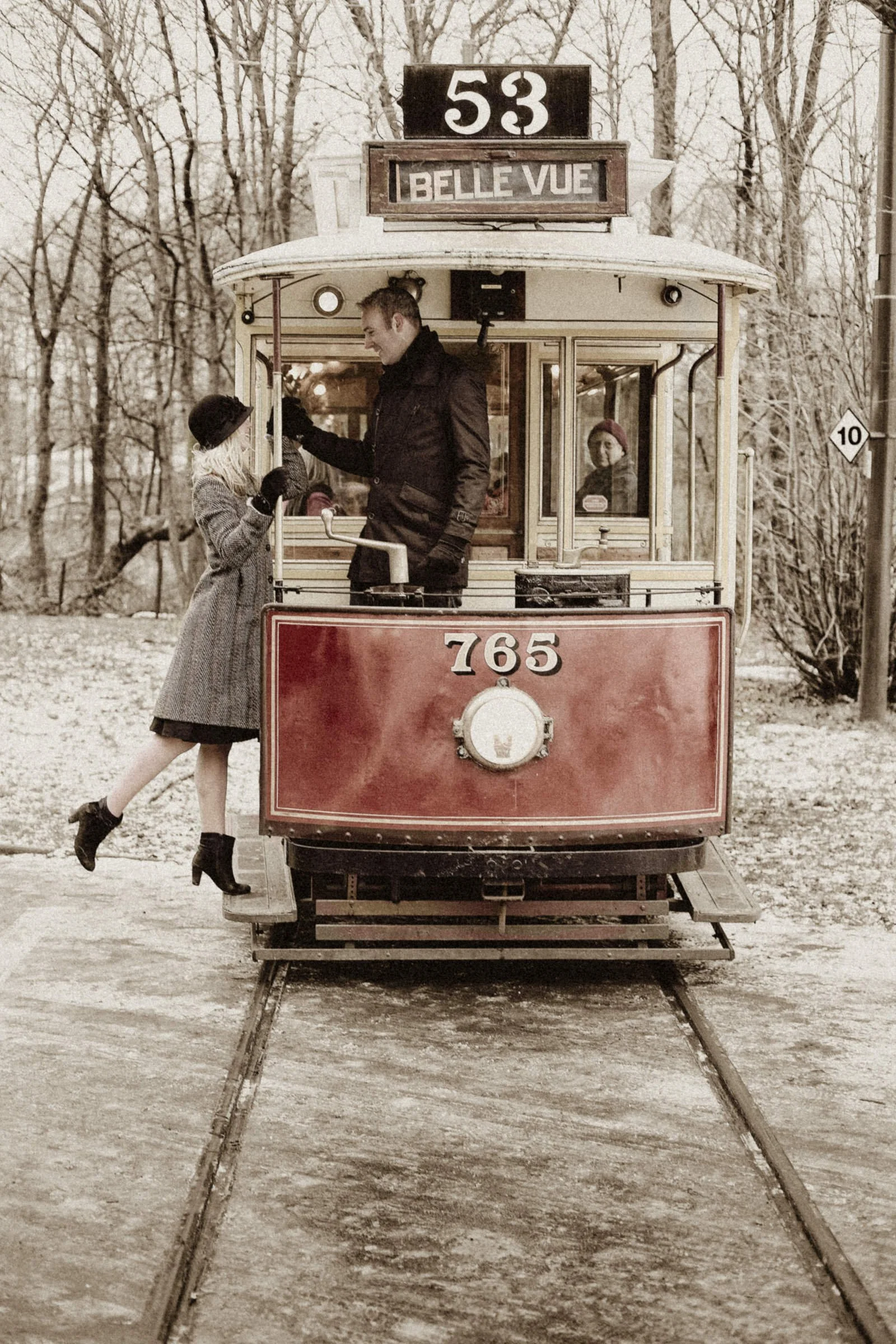 A vintage red trolley with the route number 53 and destination 'Belle Vue' in a snowy park. A man in black and a young girl in a coat and hat are interacting on the trolley, while another woman looks through the window behind them.