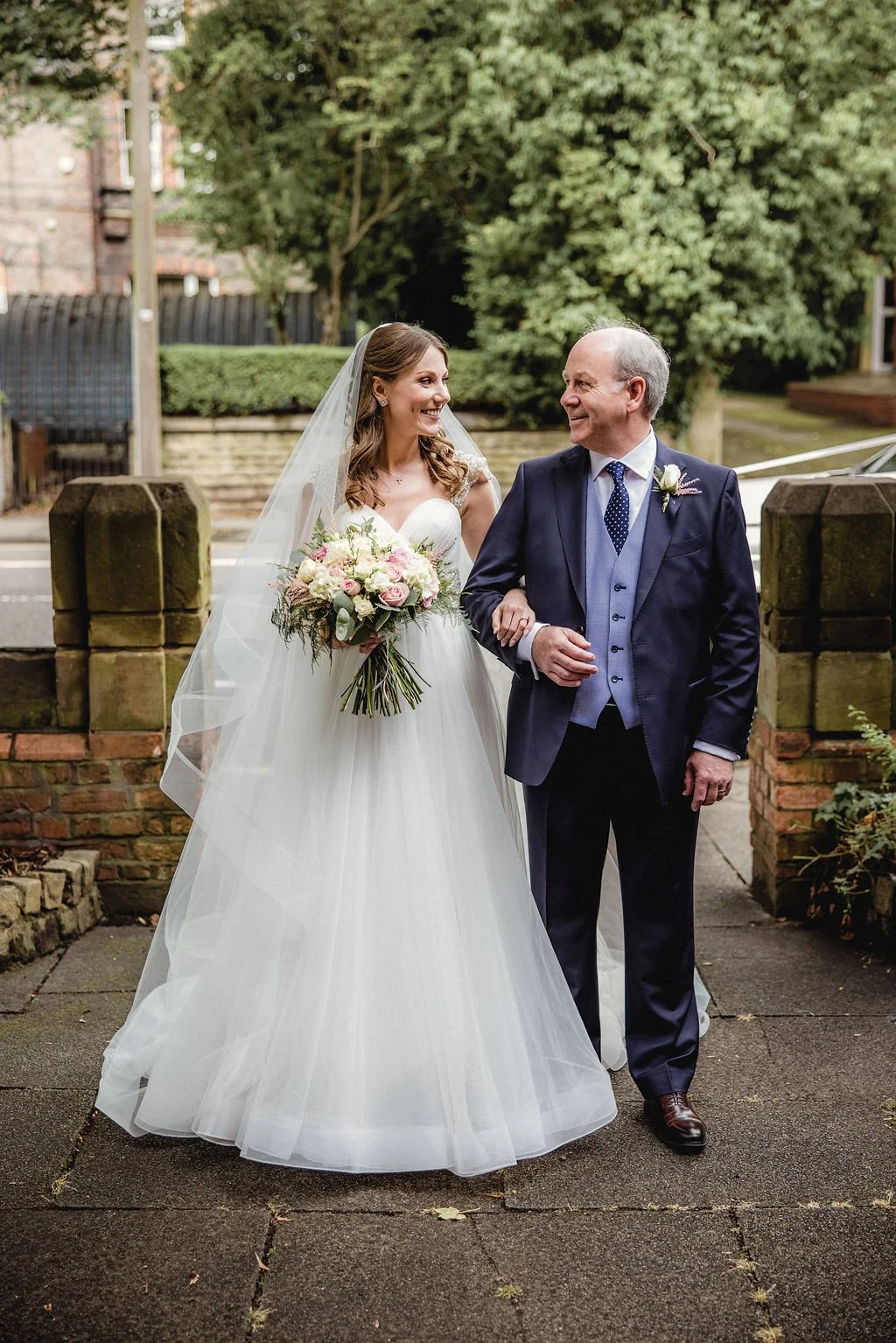 A bride in a white wedding gown holding a bouquet of pink and white flowers, walking arm-in-arm with a smiling man in a dark blue suit, outdoors near a brick wall and lush greenery.