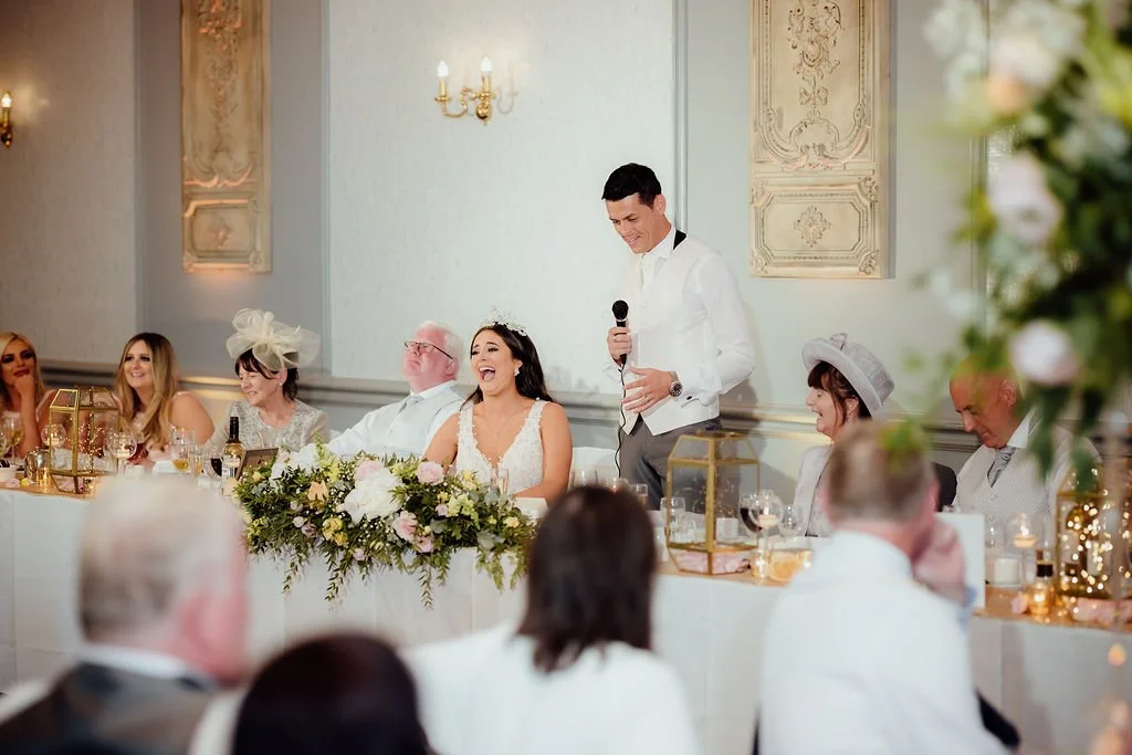 Wedding reception with a bride and groom seated at a decorated table, a man giving a speech must be the best man, guests seated on each side with drinks, flowers, and decorations, elegant interior with ornate wall art, soft lighting.