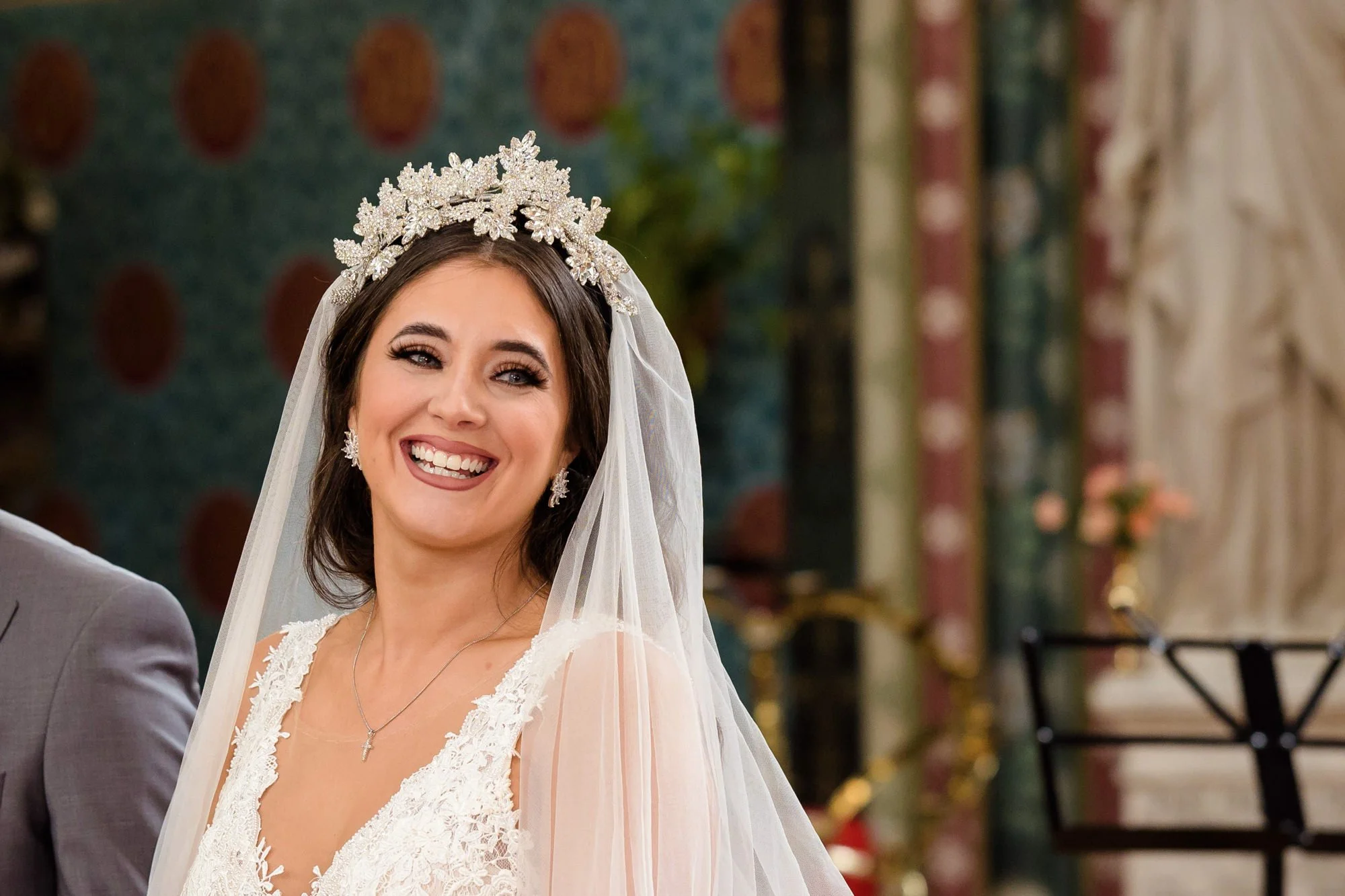 A smiling bride wearing a lace wedding dress, a silver tiara with floral designs, and a veil, standing indoors with ornate decor in the background.
