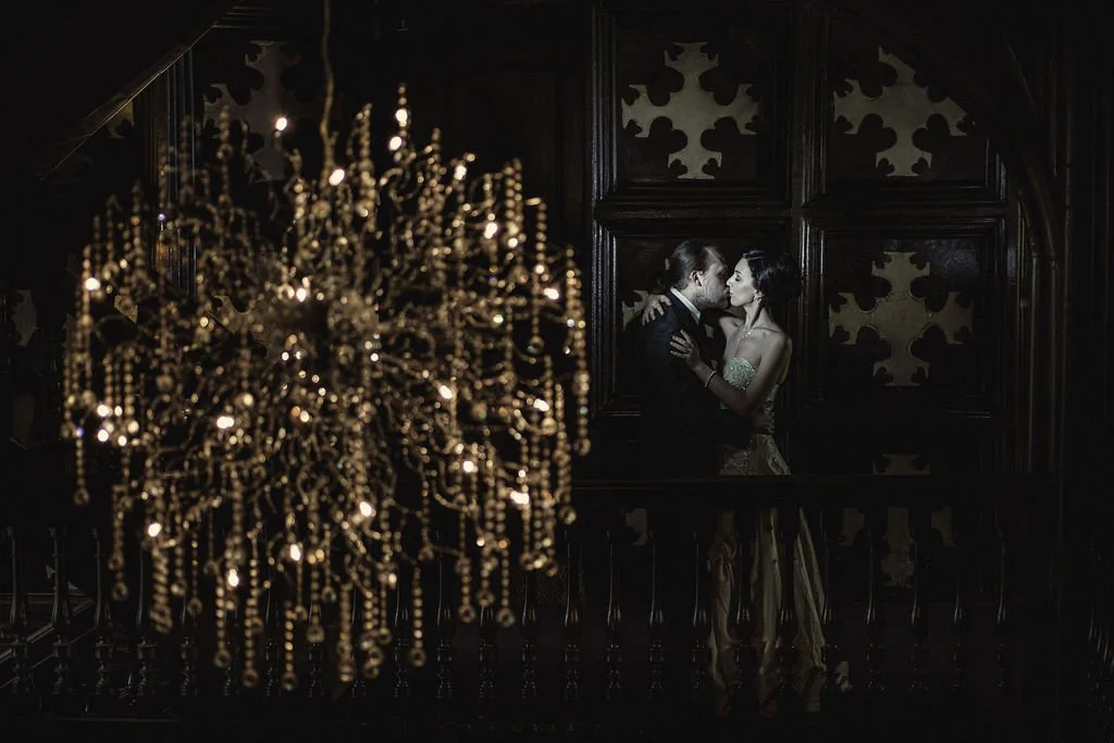 A couple dancing closely in a dimly lit, ornate room with a large, gold chandelier in the foreground.