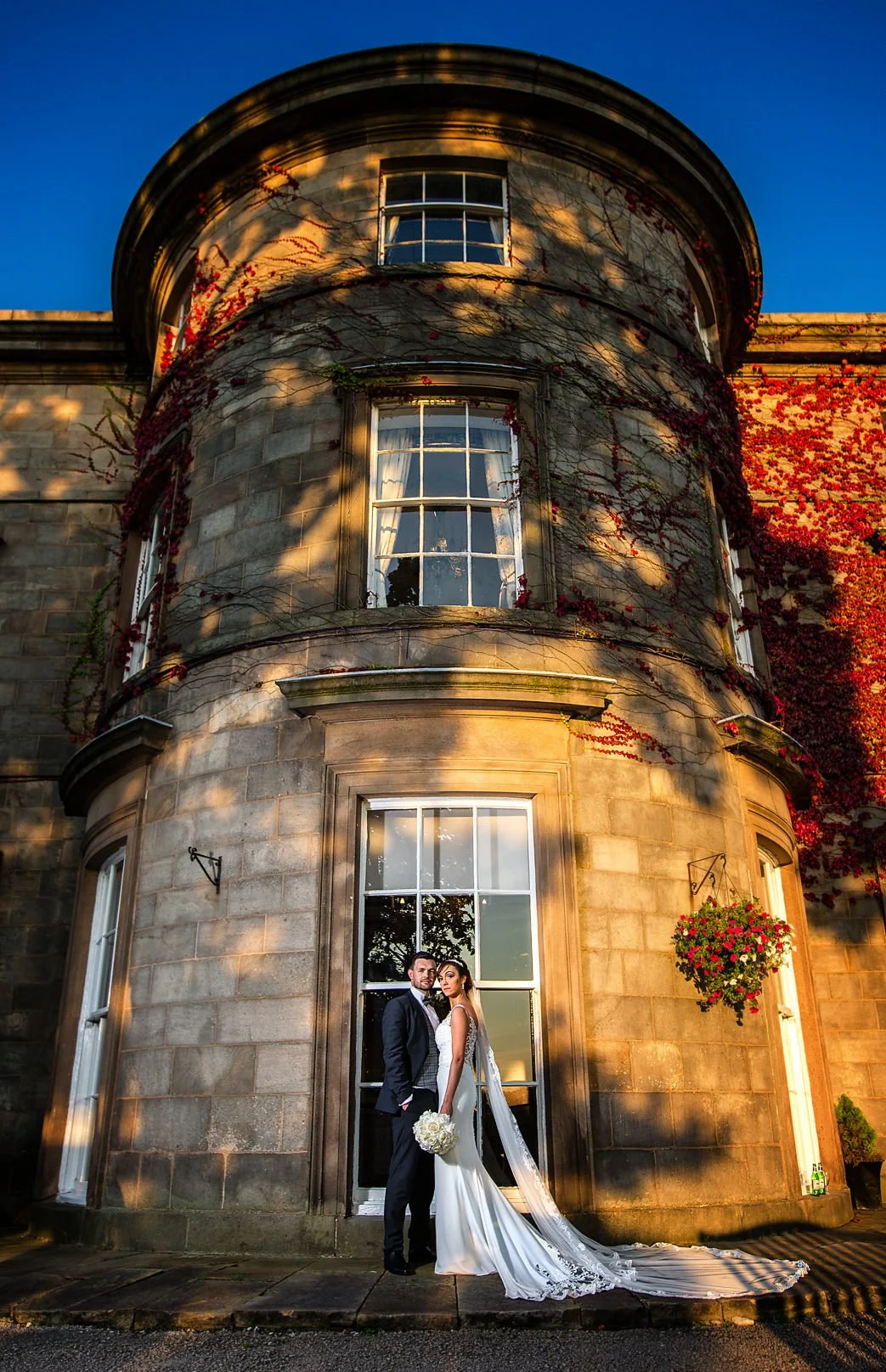 A newlywed couple stands in front of a historic stone building with large windows, illuminated by warm sunset light. The bride wears a white wedding gown with a long train and veil, holding a bouquet of white flowers. The groom wears a dark suit and 
