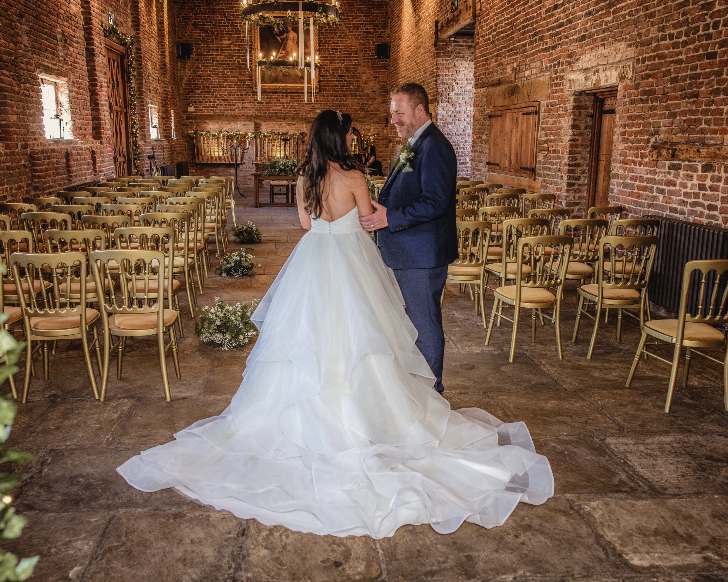 A bride and groom holding hands and smiling at each other inside a rustic brick wedding venue with gold chairs.