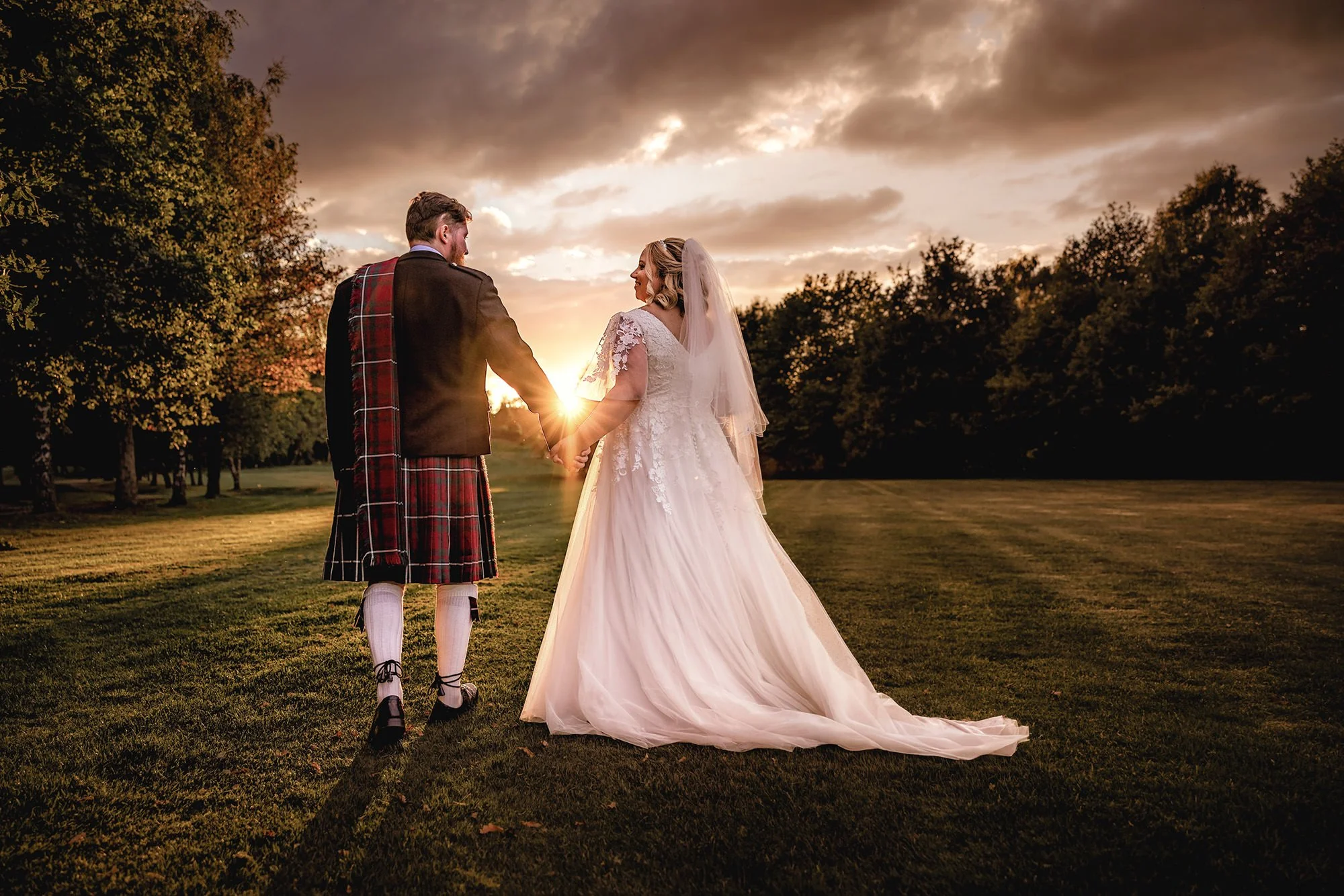 A bride and groom holding hands at sunset in a park. The groom wears a traditional Scottish kilt and jacket, and the bride wears a white wedding gown and veil. Trees line the background with a cloudy sky illuminated by the setting sun.