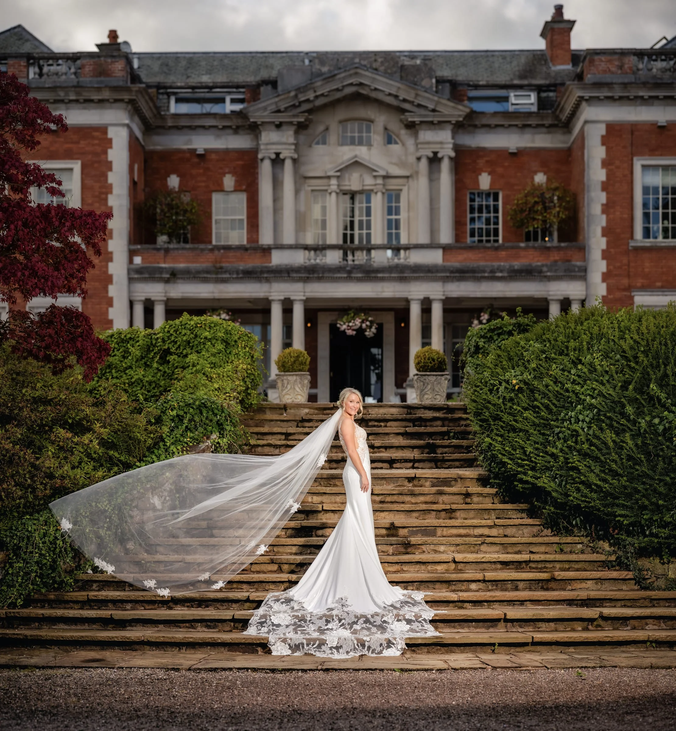 Bride standing on stone steps in front of a large historic mansion, wearing a white wedding gown with a long train and veil.