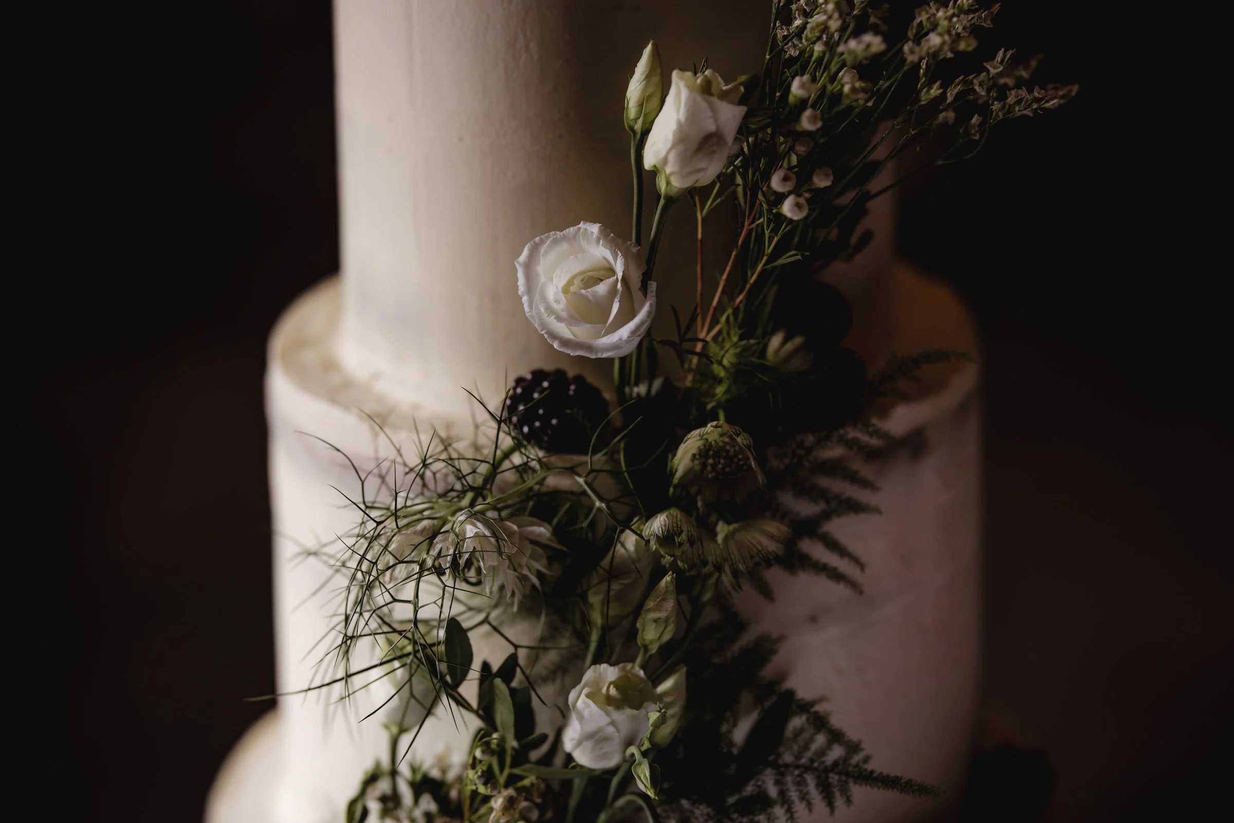 Close-up of a white wedding cake decorated with white roses, greenery, and dark berries.