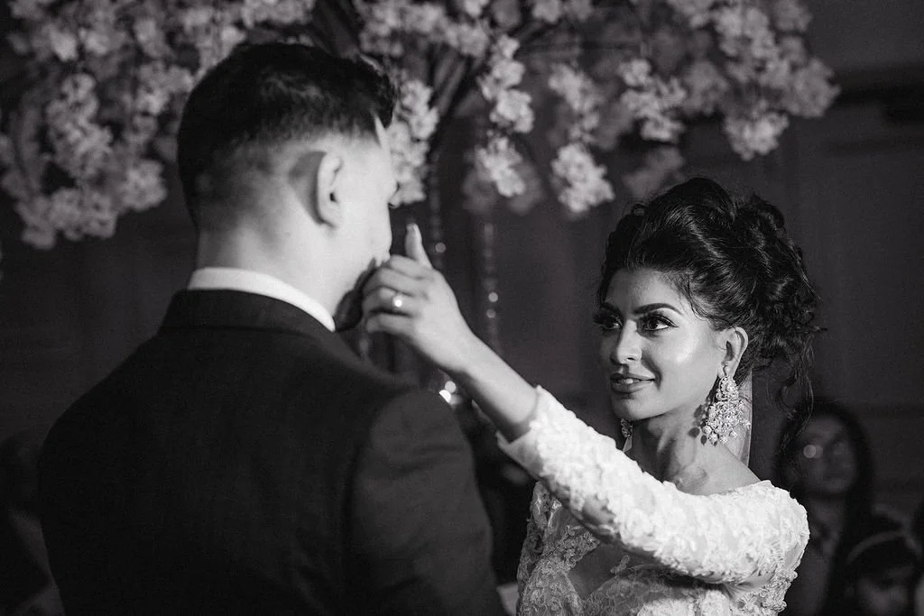 A woman with dark, curly hair and large earrings is touching a man's face during a dance, with flowers in the background.
