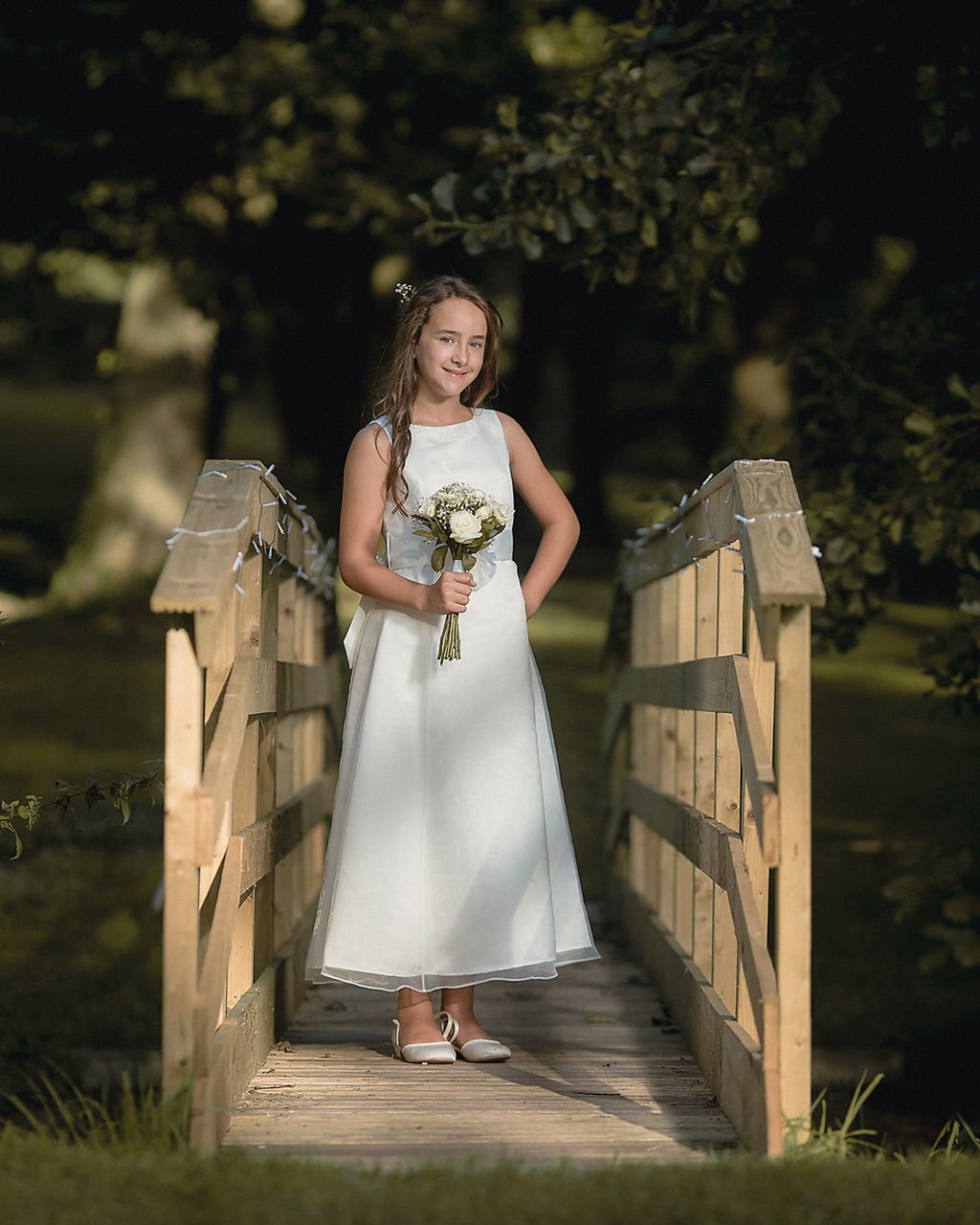 A young girl in a white dress holding a bouquet standing on a wooden bridge at night, surrounded by trees.