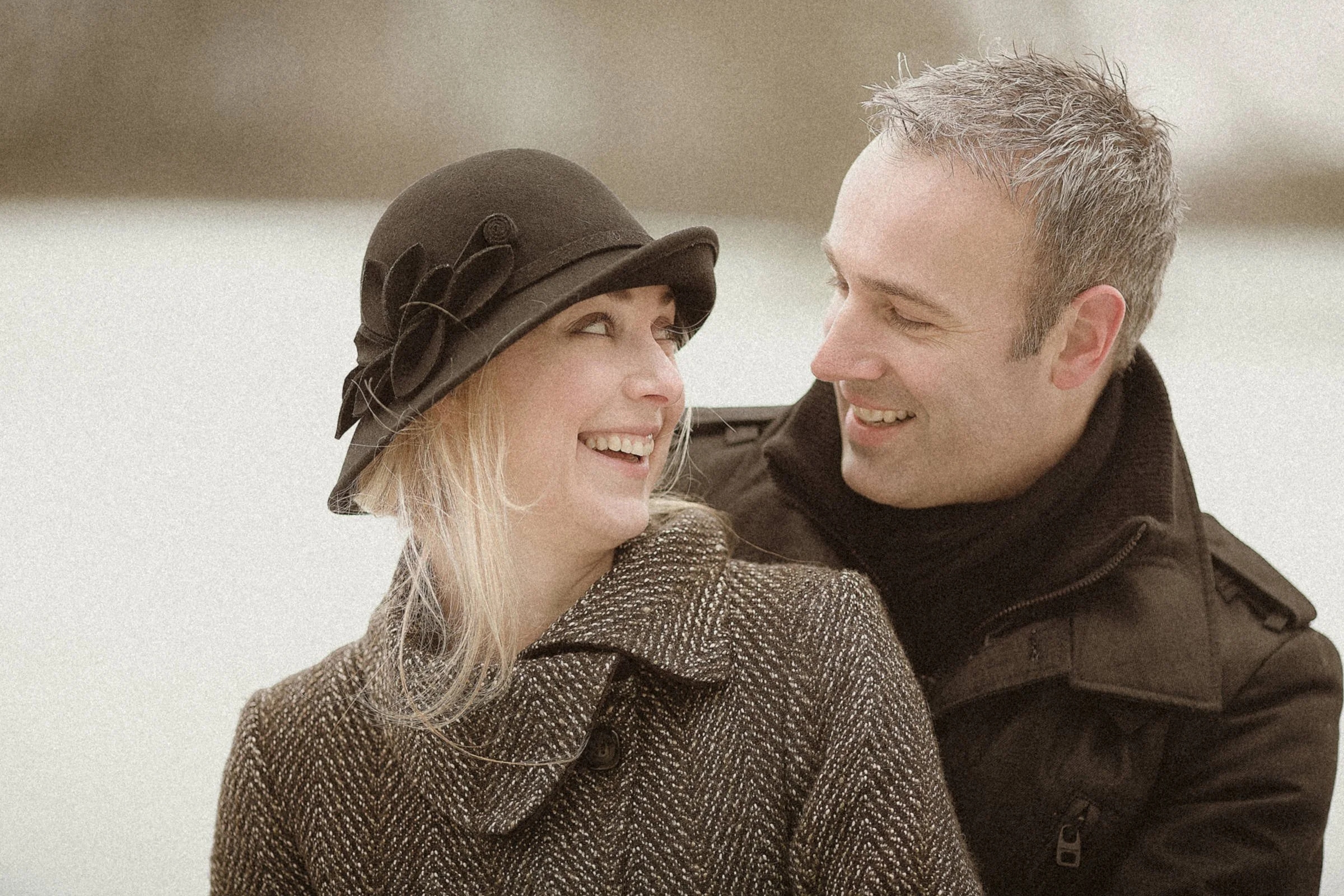 A man and a woman smiling at each other, outdoor setting, woman wearing a dark hat with decorative flower and brown coat, man in a dark jacket, both cheerful and close together.