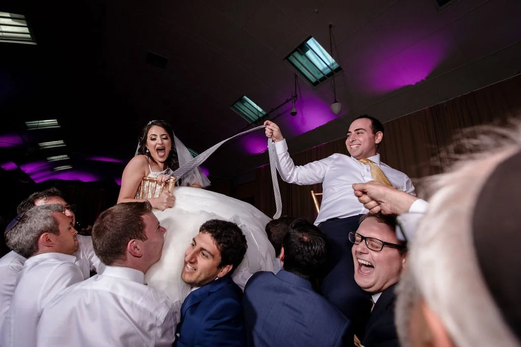 Bride and groom being lifted on a chair at a wedding celebration, surrounded by guests, with everyone smiling and laughing in a festive indoor setting with purple lighting.