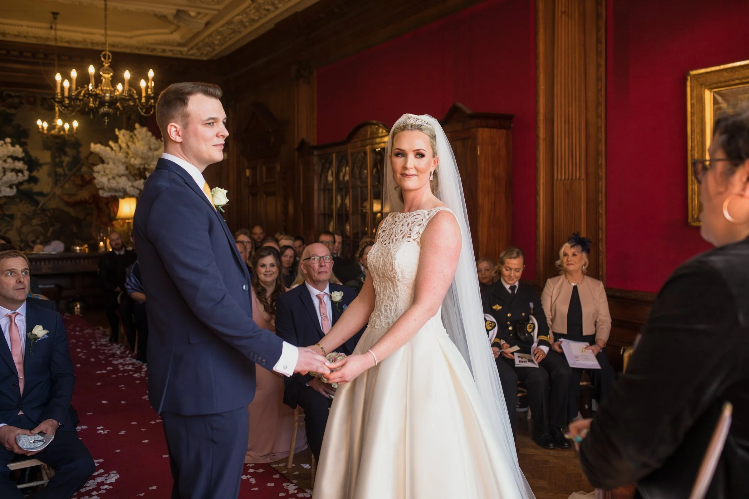 A bride and groom holding hands during their wedding ceremony in an elegant room with red walls and ornate woodwork, surrounded by seated guests.
