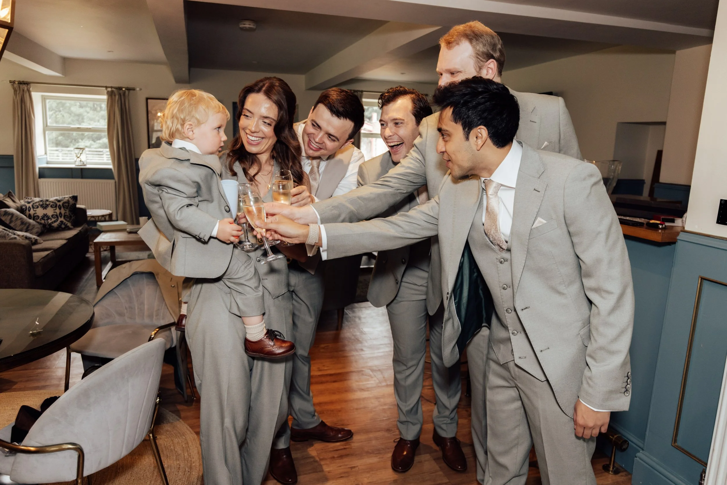 A group of six people, five men and one woman, in suits, and one small boy in a gray suit, celebrating with champagne in a living room with beige and blue decor.