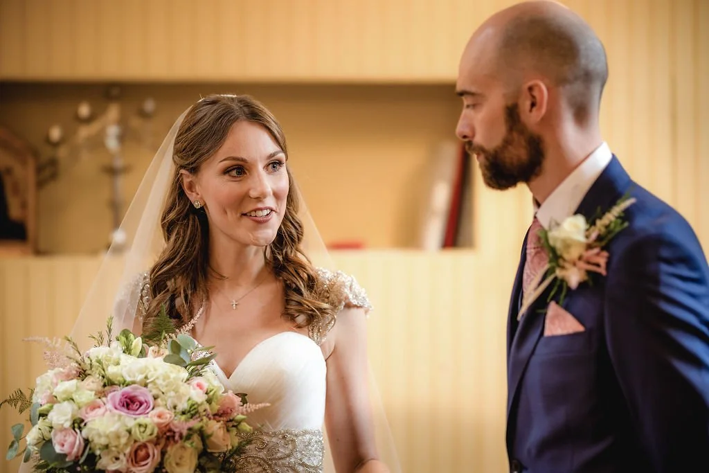 A bride and groom during their wedding ceremony indoors, with they facing each other and the bride holding a bouquet of pink and white flowers.