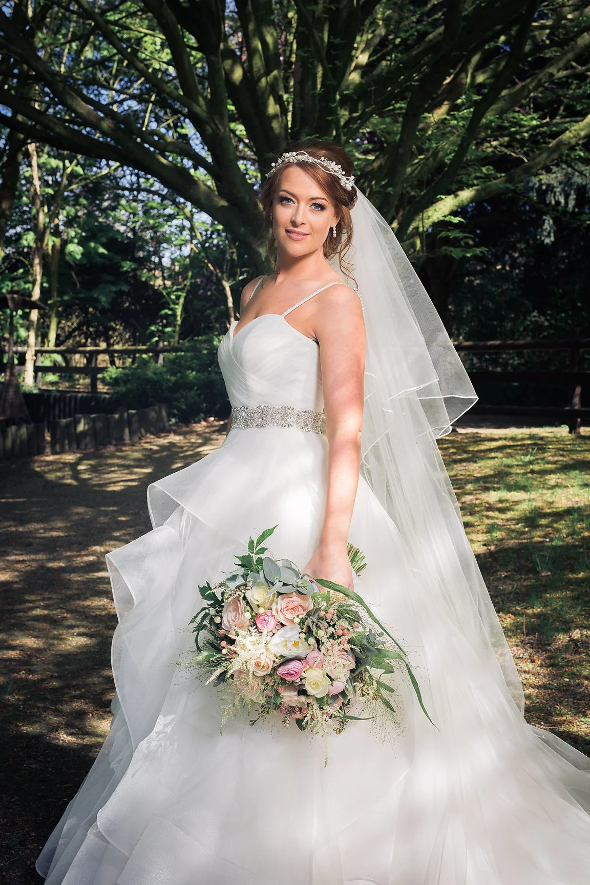 A bride in a white wedding gown holding a bouquet of pink and white roses, standing outdoors in a wooded area with sunlight filtering through the trees.