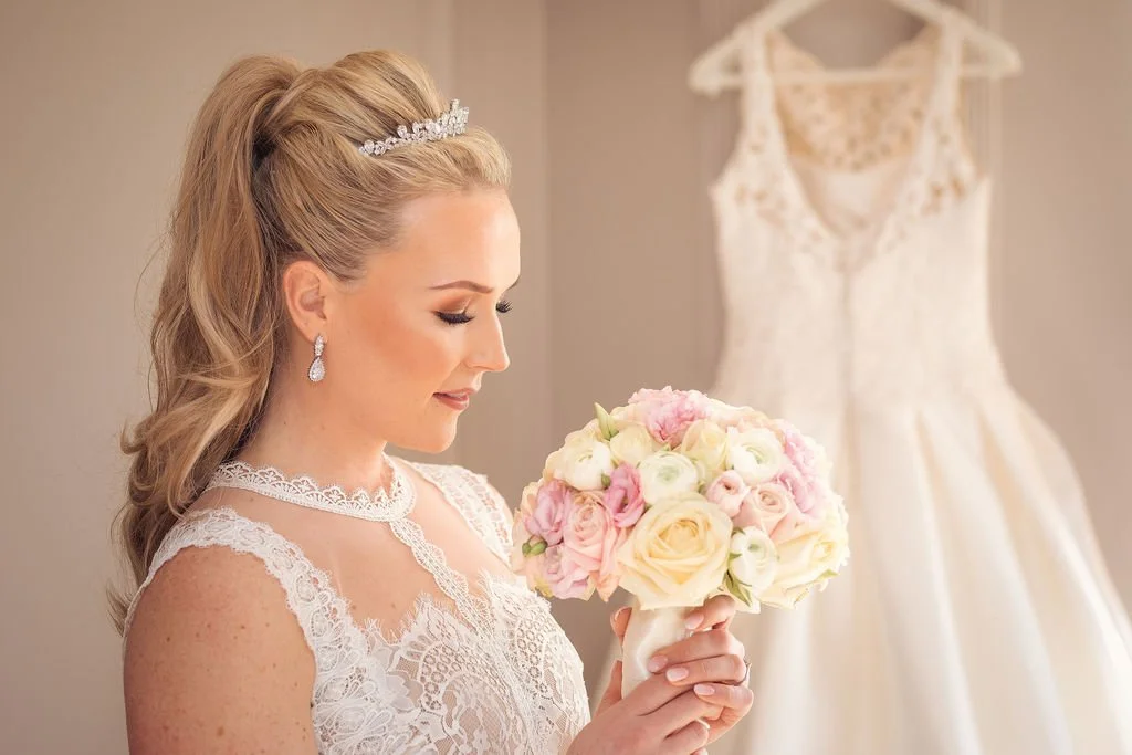 Blonde haired bride in white attire looks at her pink, yellow and white bouquet of flowers with her white wedding dress hung up in the background.