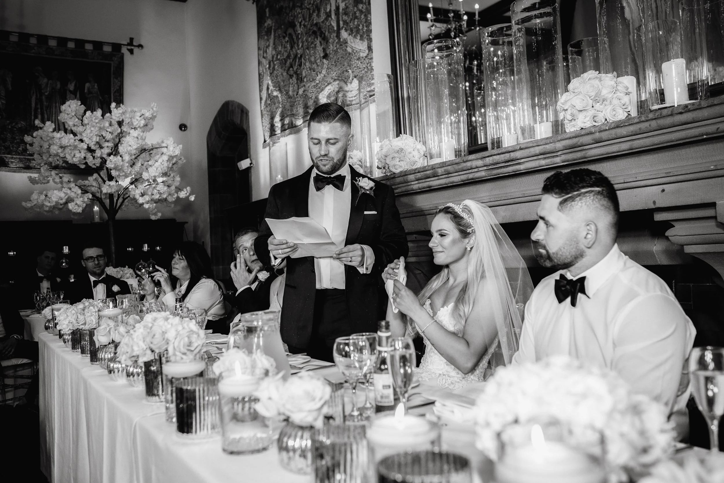 A black and white photo of a wedding reception, with a man in a tuxedo giving a speech while the bride and groom sit at the head table among guests. The bride wears a veil and the groom is also in tuxedo.