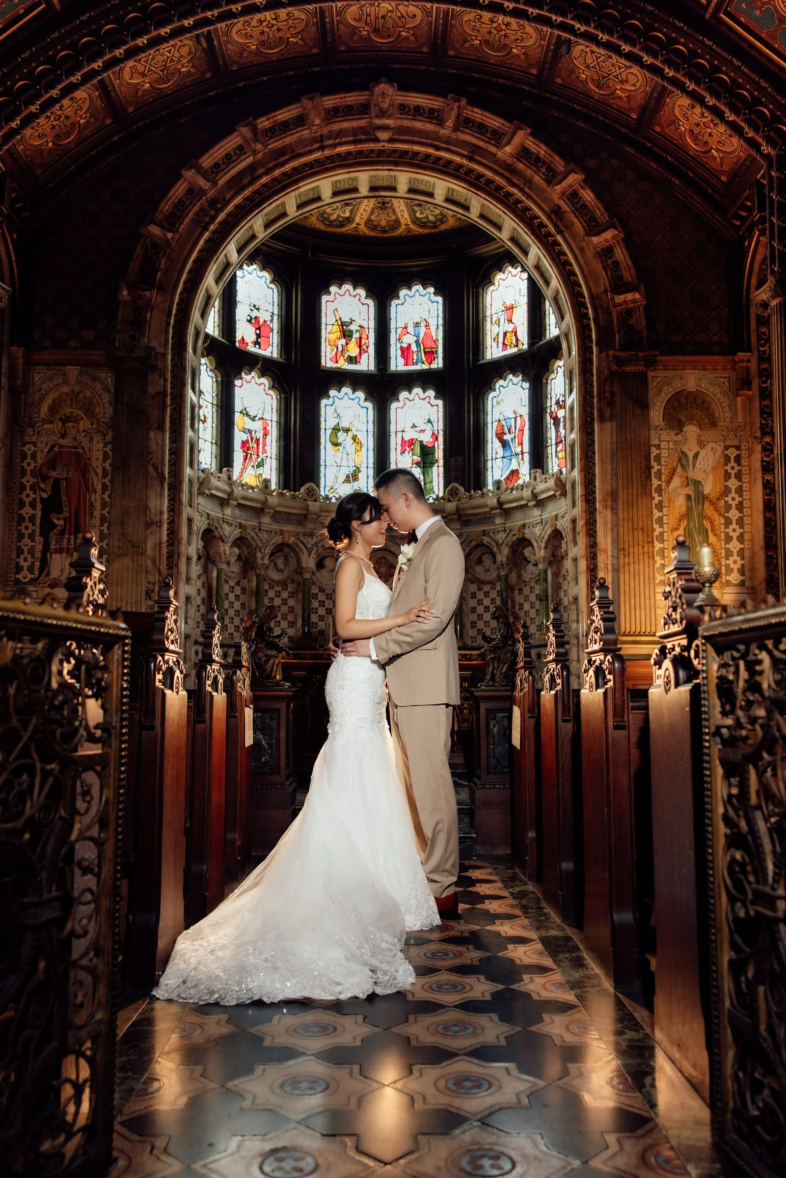 A bride and groom stand close together inside a church with stained glass windows, ornate woodwork, and decorative tile floor, as they touch foreheads and hold each other during their wedding ceremony.