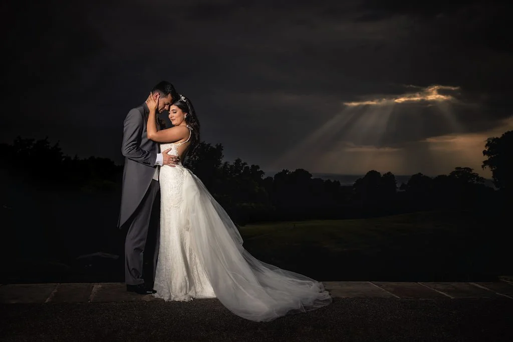 A bride and groom standing close together outdoors at sunset, with the groom in a suit and the bride in a wedding gown and veil, embracing as dark clouds and rays of sunlight are in the sky.