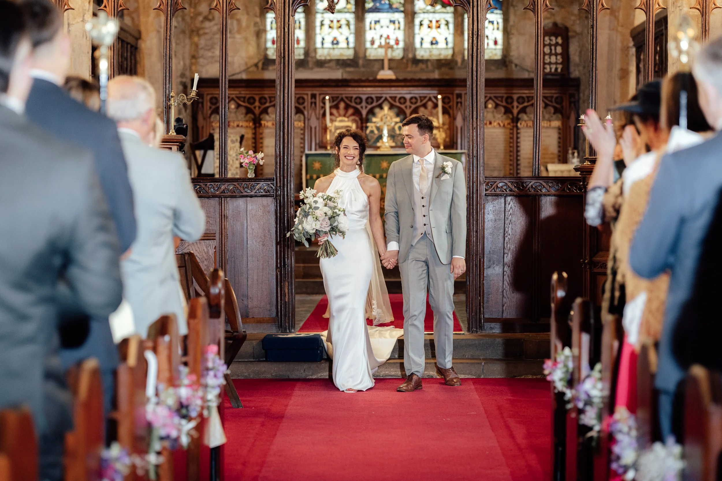 A bride and groom walking hand in hand down the aisle during their wedding ceremony inside a church, with guests seated on either side watching and taking photos.