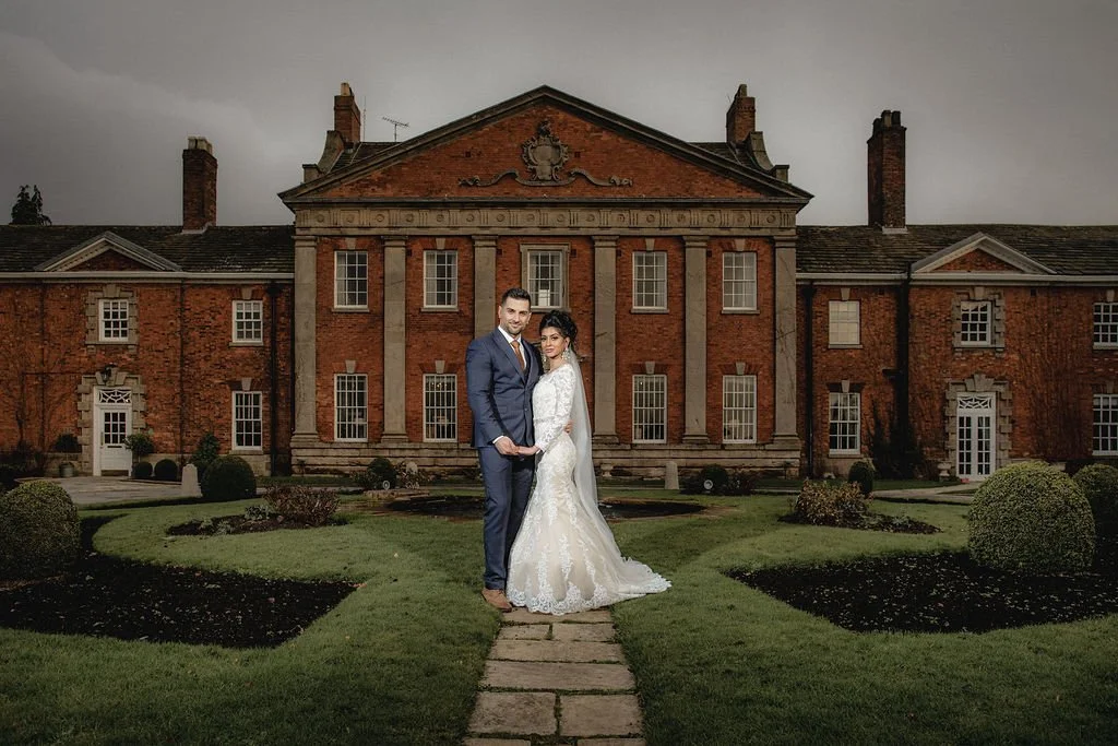 A bride and groom standing on a stone pathway in front of a historic brick mansion with a well-manicured lawn and shrubs, during overcast weather.
