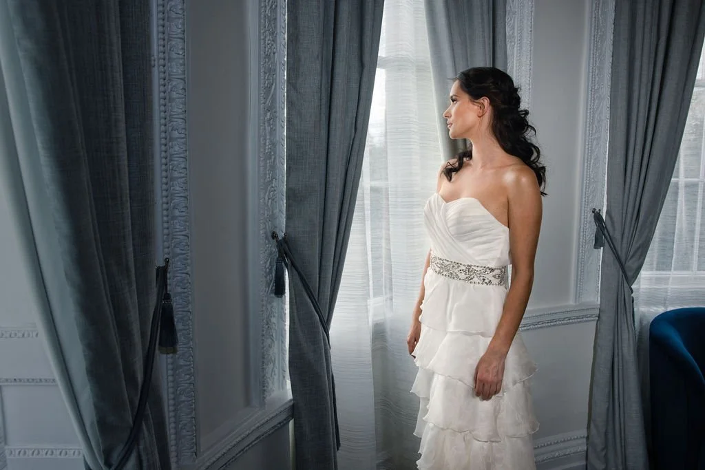 A woman in a strapless white wedding gown standing by a window with blue curtains, looking outside.