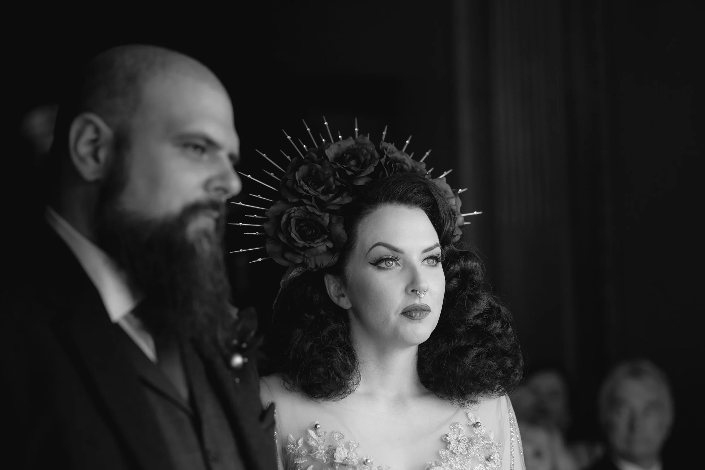 Black and white photo of a woman with dark, curly hair wearing a flower crown and regal gown, with a man in the foreground with a beard and bald head, both at a formal event.