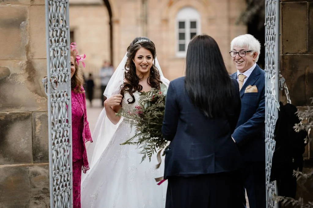 A bride smiling at a woman holding a bouquet, standing outside a church with a man in a suit, all engaged in conversation.