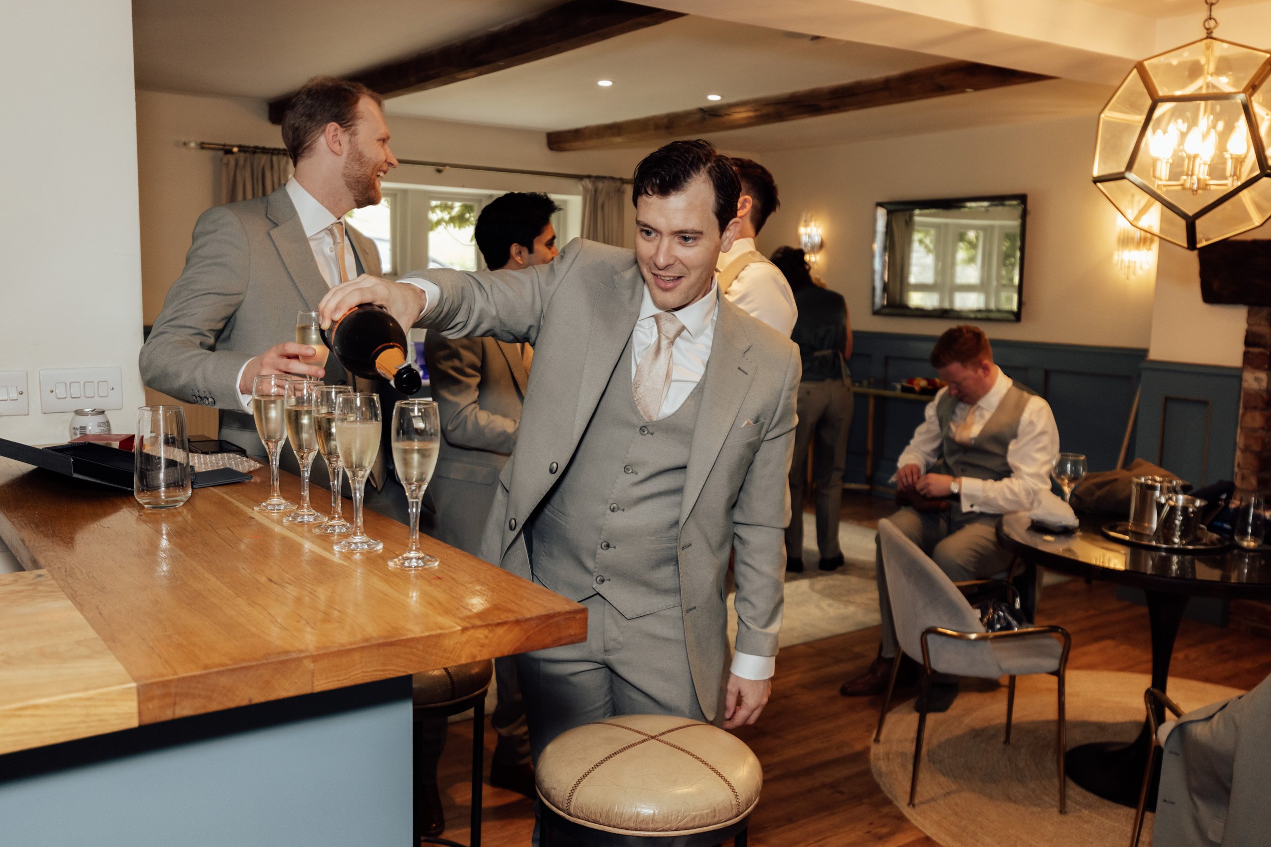 A man in a gray suit pouring champagne into glasses at a celebration event in a cozy, warmly lit room with wooden beams, large windows, and other guests in formal attire.
