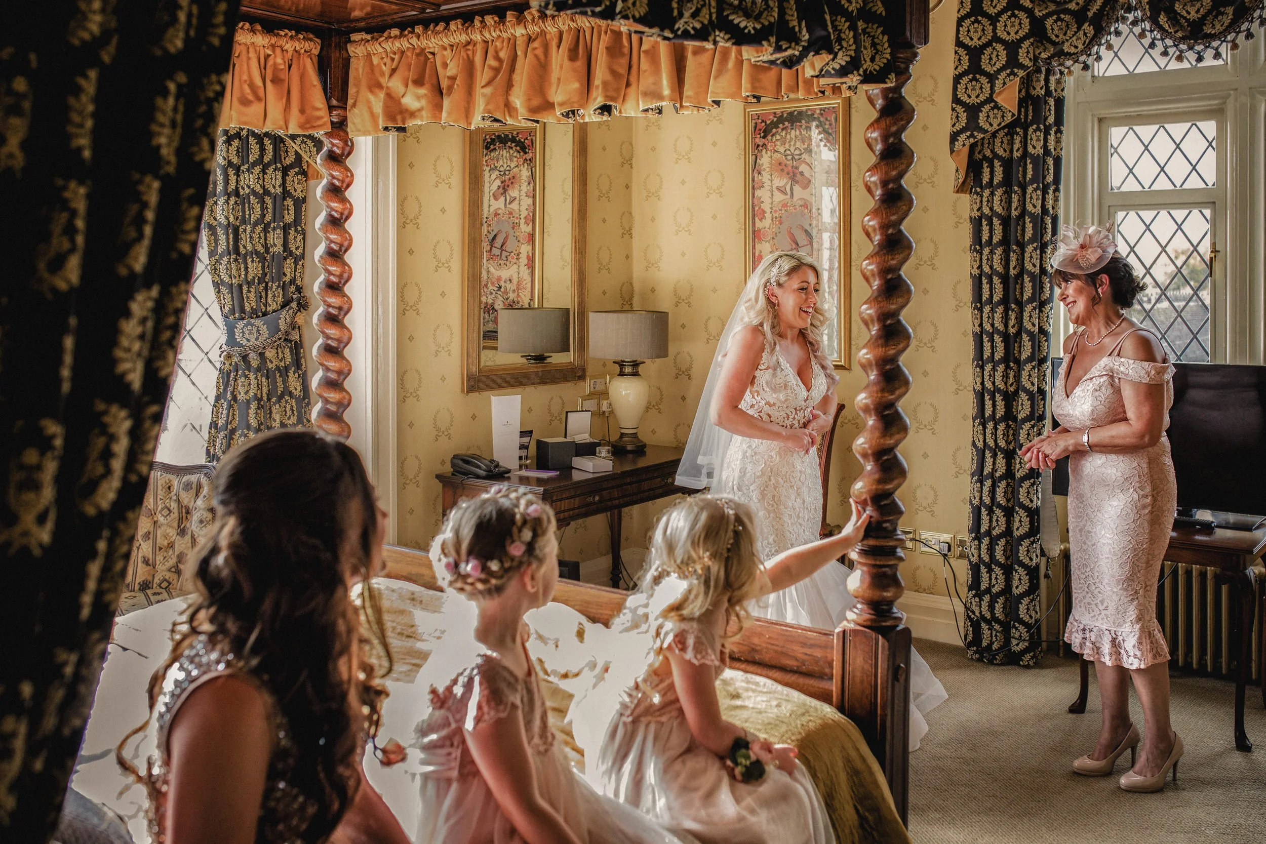 A bride in a wedding dress and veil standing in a room with four young girls sitting on a bed, with one girl reaching towards a woman in a pink dress, who is standing near a window. The room has ornate curtains, framed artwork, and a dresser with lam