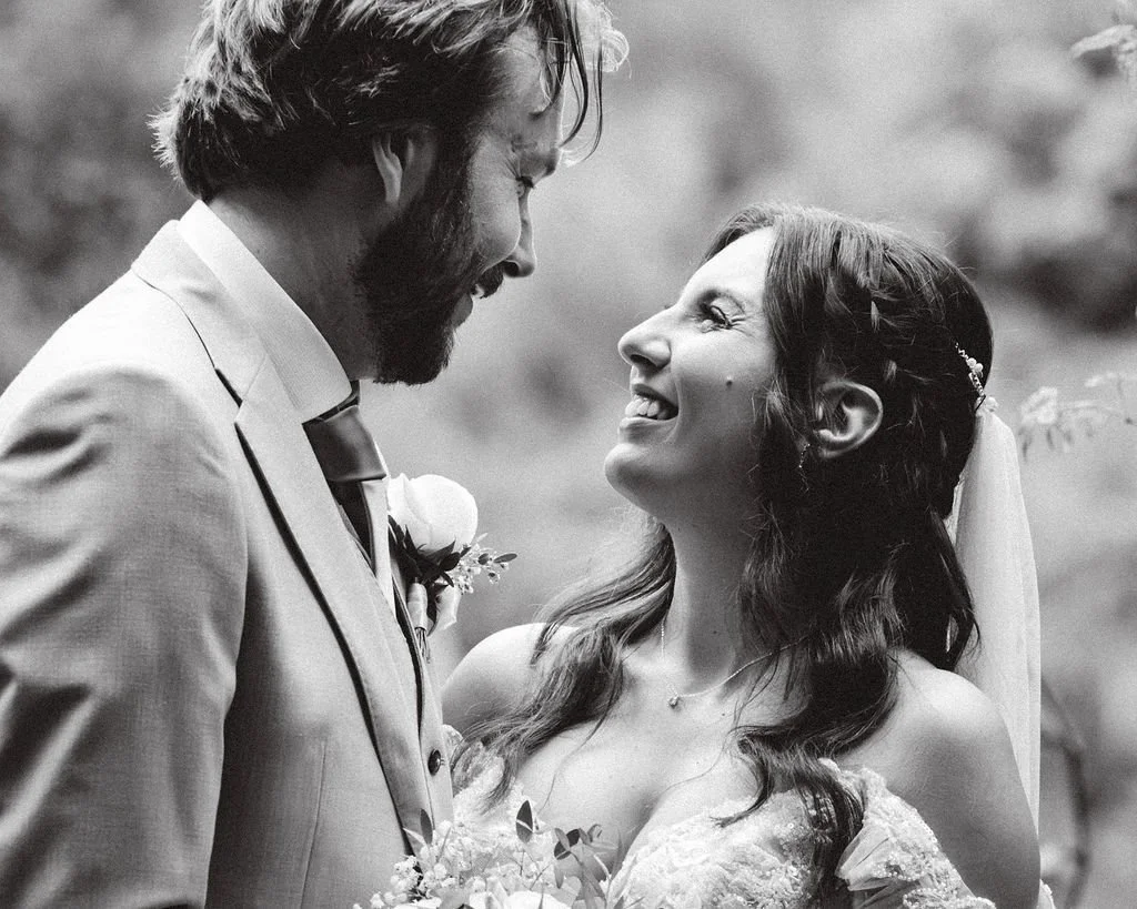 A black and white photo of a bride and groom smiling at each other, standing close with faces near each other, outdoors.