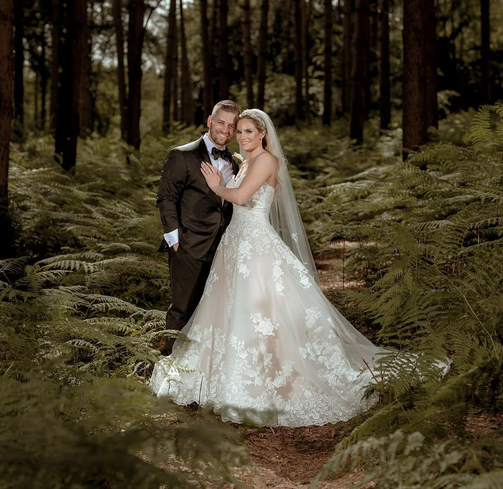 A newlywed couple in wedding attire standing in a forest with tall trees and ferns, smiling and holding each other.