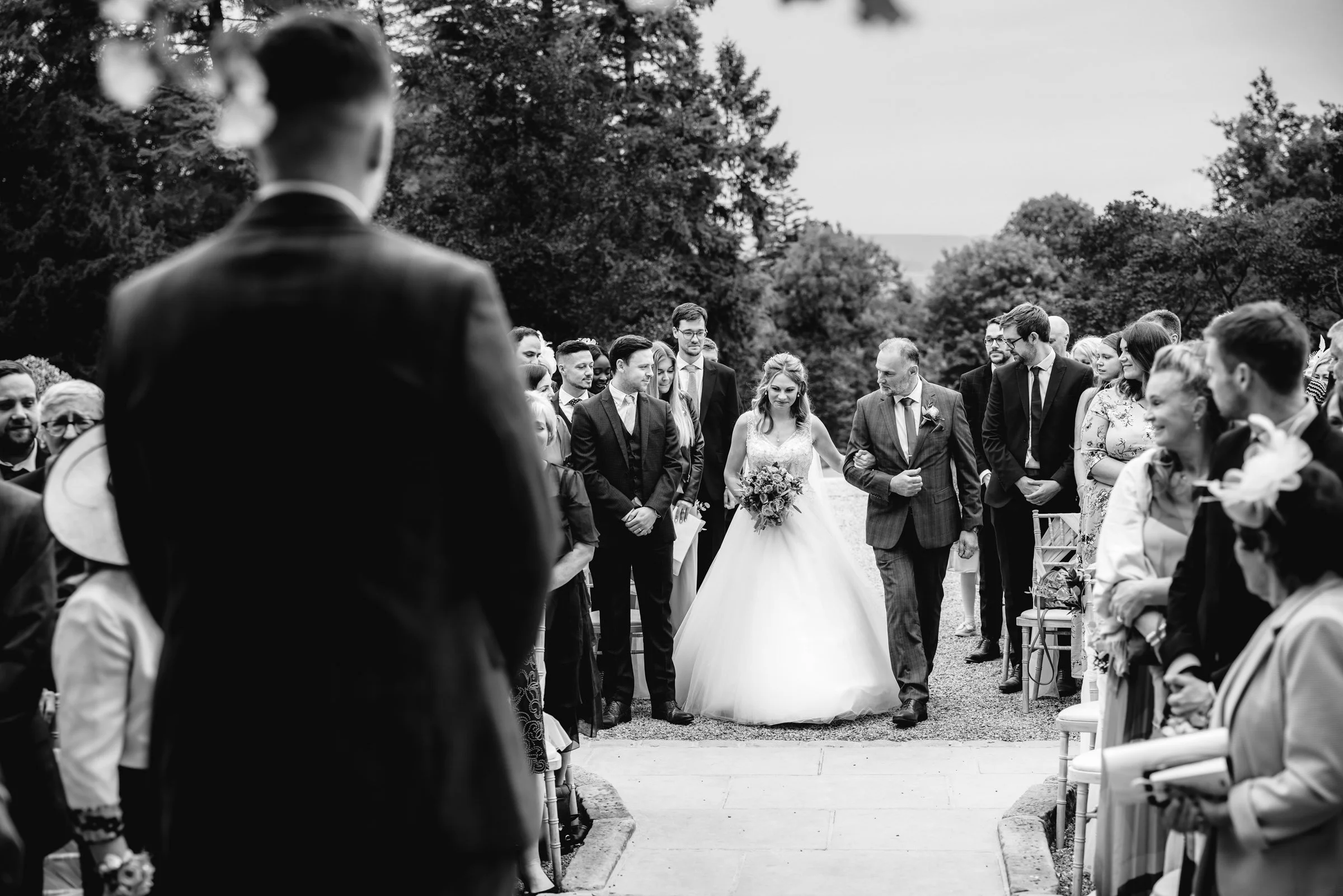 A black and white photo of a wedding ceremony outdoors. The bride in a white gown is walking down the aisle, accompanied by an older man. Guests stand on either side, watching her. The scene is surrounded by trees.