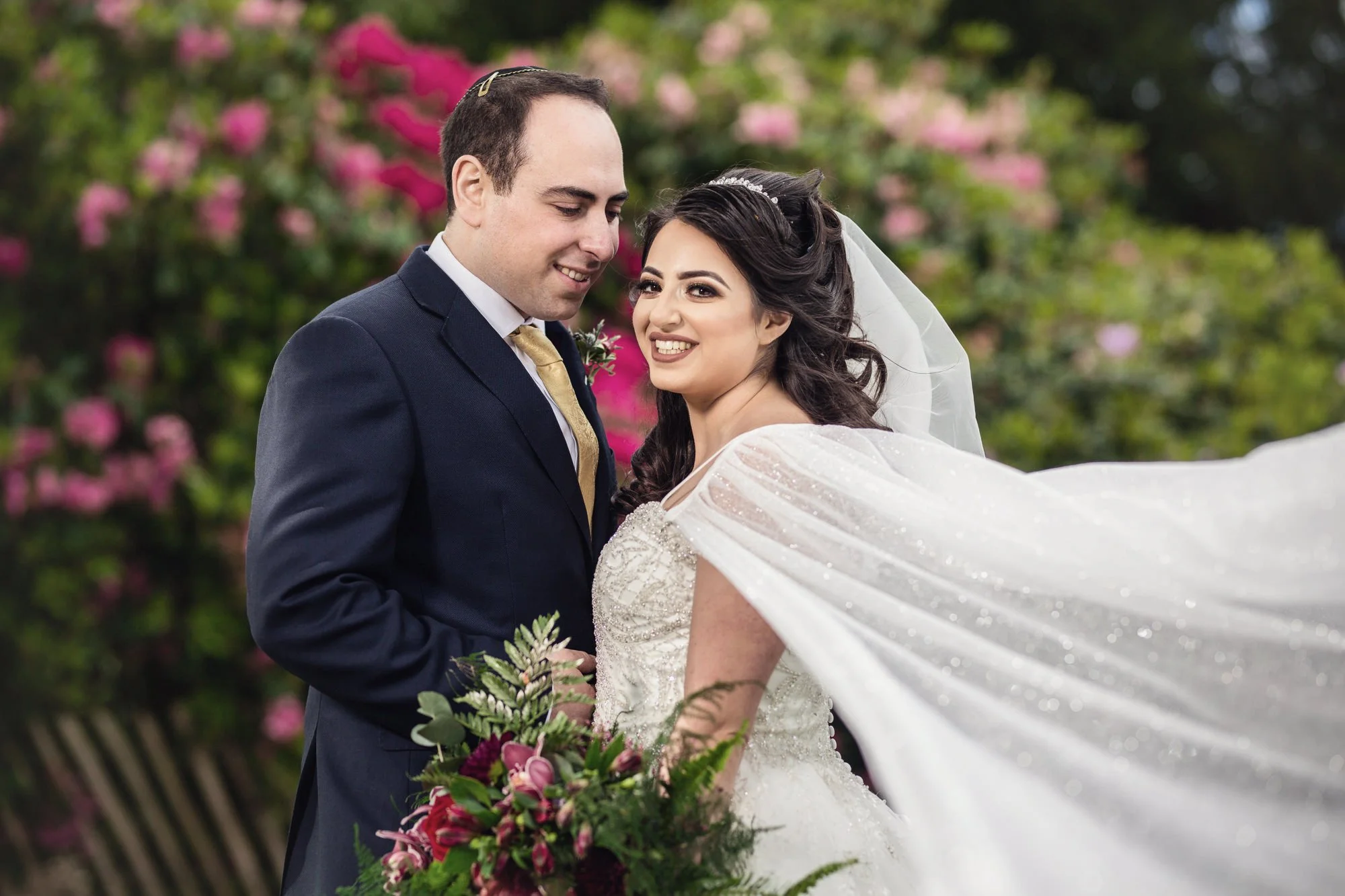 A bride and groom on their wedding day, outdoors with pink flowers in the background. The groom is wearing a dark suit with a gold tie, and the bride is in a white wedding gown with a veil, holding a bouquet of flowers.